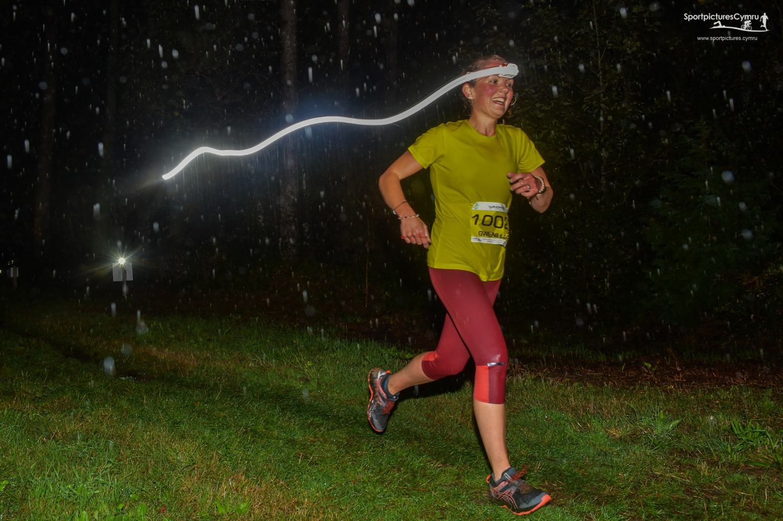 A person wearing a bright yellow shirt, red leggings, and a headlamp runs through the rain at night. The headlamp creates a streak of light above their head. The background is dark and forested, illuminated only by the headlamp and distant lights.