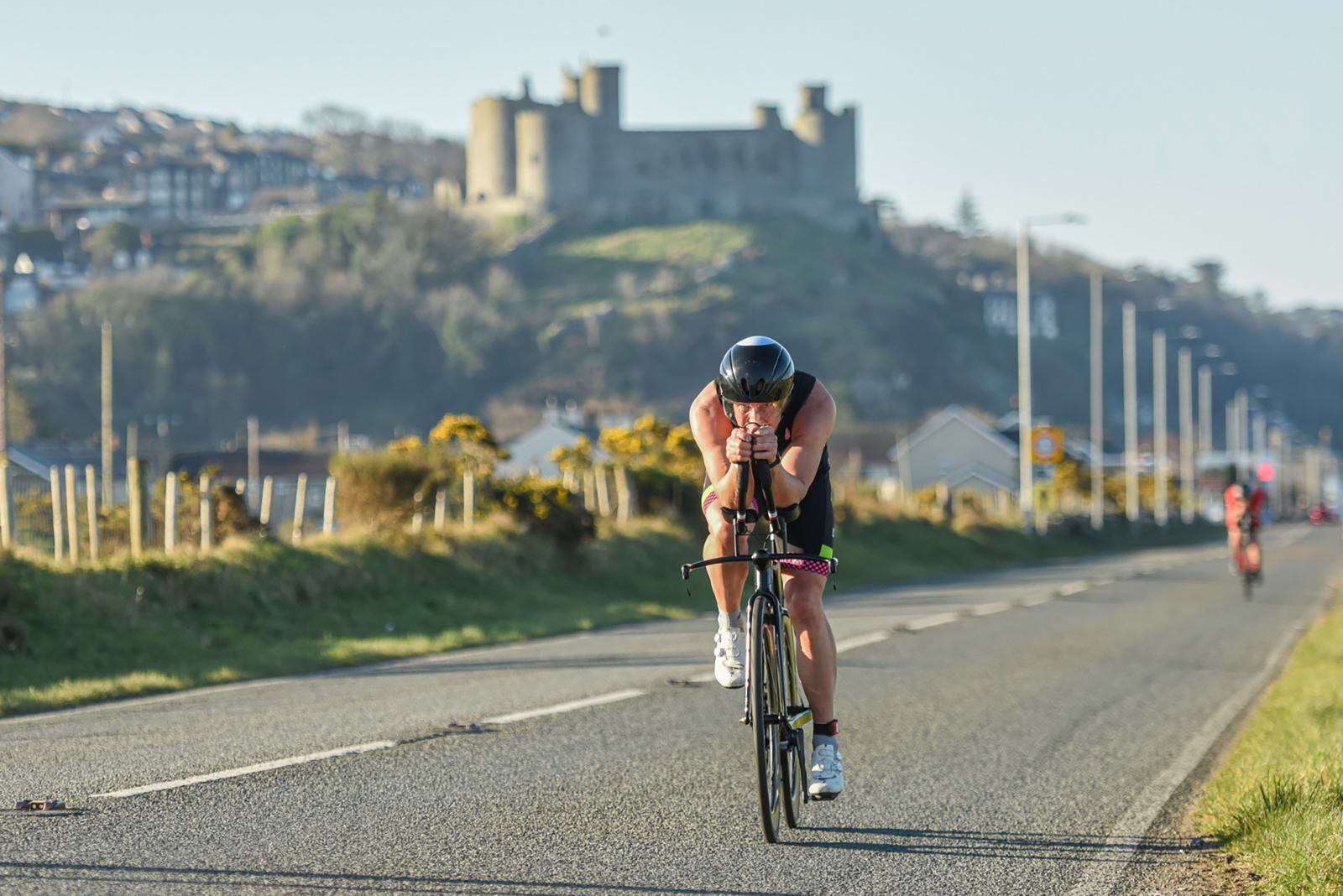 A cyclist in a black helmet and sports attire rides on a road, leaning forward on the handlebars. The background features a large, historic castle atop a hill, with town buildings and trees surrounding it. The sky is clear and blue.