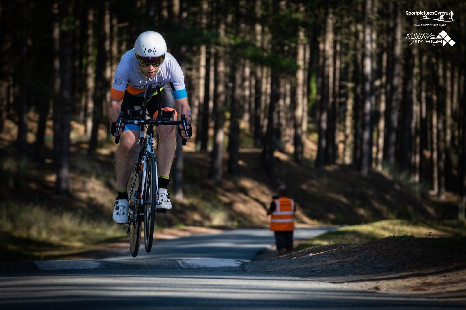 A cyclist in racing gear and a white helmet rides a bicycle on a wooded road during a race. Another person in an orange vest stands further down the road, facing away. Sunlight filters through the trees, creating dappled shadows on the path.