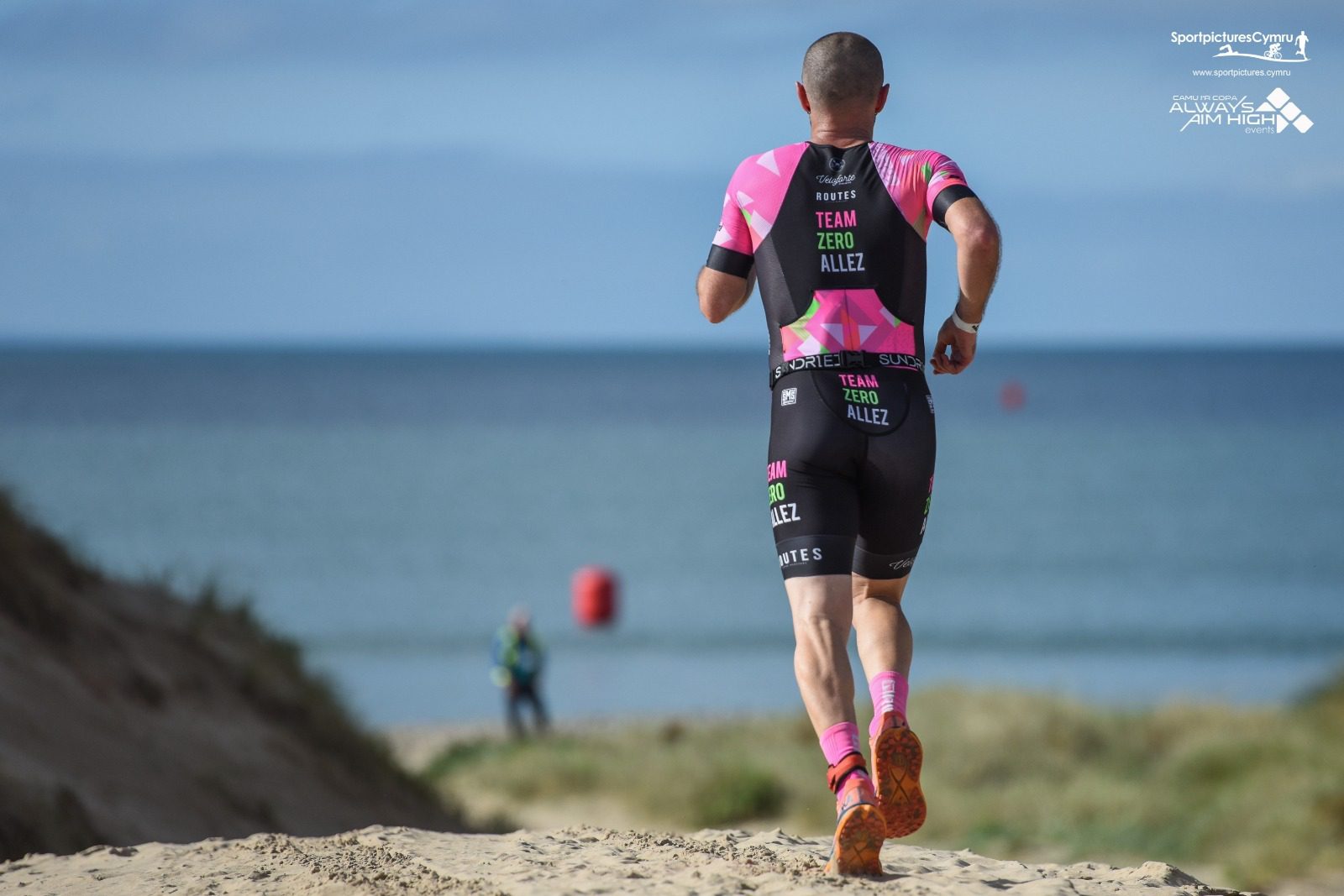 A triathlete in a pink and black uniform with the words "Team Zero Allez" on the back is running on a sandy beach towards the ocean. There are red buoys in the water and a blue sky in the background. Another person can be seen in the distance near the water.