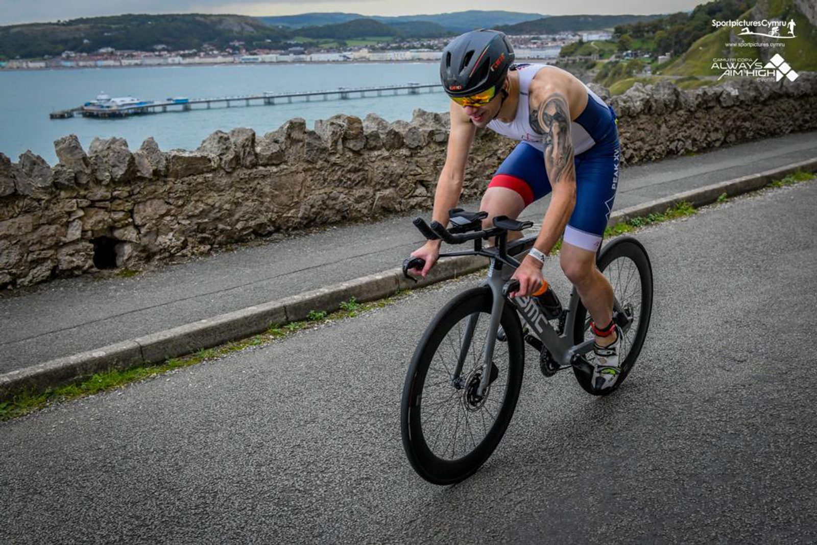 A triathlete wearing a helmet, sunglasses, and a tri-suit rides a road bike along a scenic coastal route. A stone wall separates the road from the coast. In the background, there are hills, a pier, and buildings along the shoreline. The sky is partly cloudy.