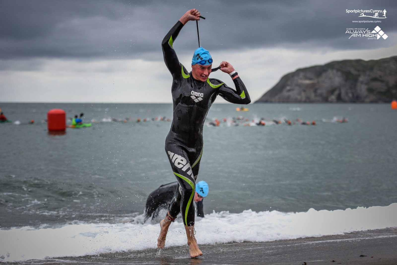 A triathlete emerging from the water in a wetsuit and blue swim cap, adjusting their goggles. They are at a beach with a rocky landscape in the background. Other swimmers are still in the water, and orange buoys mark the swim course. The sky is overcast.