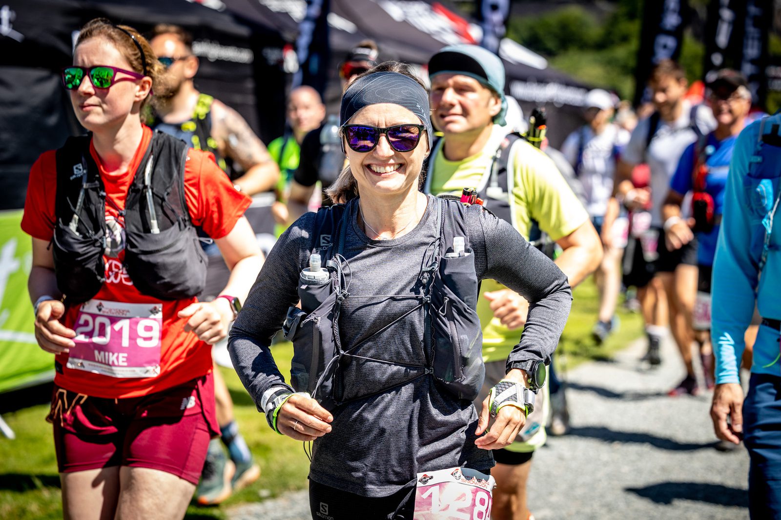 A group of runners is in the midst of a race. The woman at the center, wearing sunglasses, a headband, and a hydration vest, smiles as she runs. Other participants can be seen in the background, also wearing race bibs and gear. The sun is shining brightly.