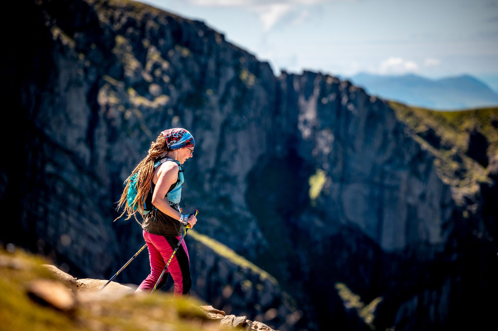 A person with long hair and a colorful bandana is hiking on a mountain trail with steep cliffs in the background. They are wearing a sleeveless shirt, red pants, and carrying hiking poles. The scene is bright with a clear blue sky.