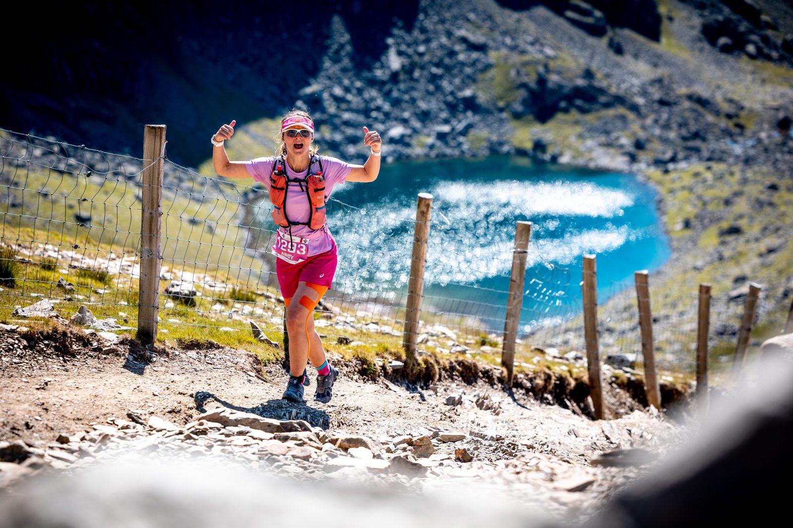 A runner dressed in pink gear and wearing a race bib number 1293 is running on a mountainous trail with a scenic view of a lake and rocky terrain in the background. The runner is giving a thumbs-up gesture with both hands.