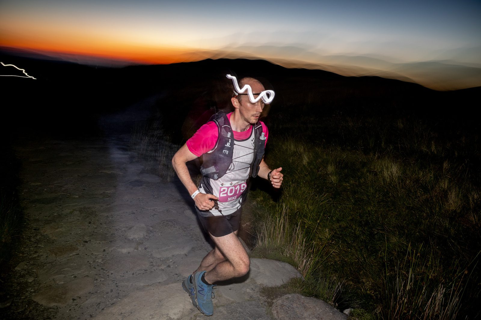 A runner wearing a headlamp and bib number 2019 jogs along a rocky path at dusk. The sky in the background displays hues of orange, pink, and blue as the sun sets, and blurred motion effects highlight the runner's movement.