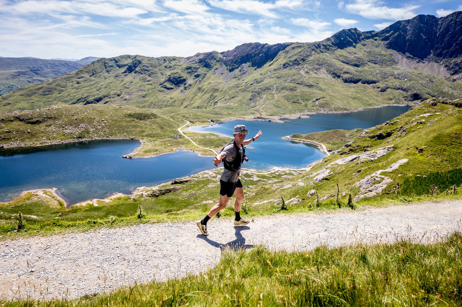 A person runs on a gravel path through a mountainous landscape with two large lakes. The sun is shining brightly, creating vibrant green hues on the hills and deep blue shades in the water. The sky is dotted with thin, wispy clouds.