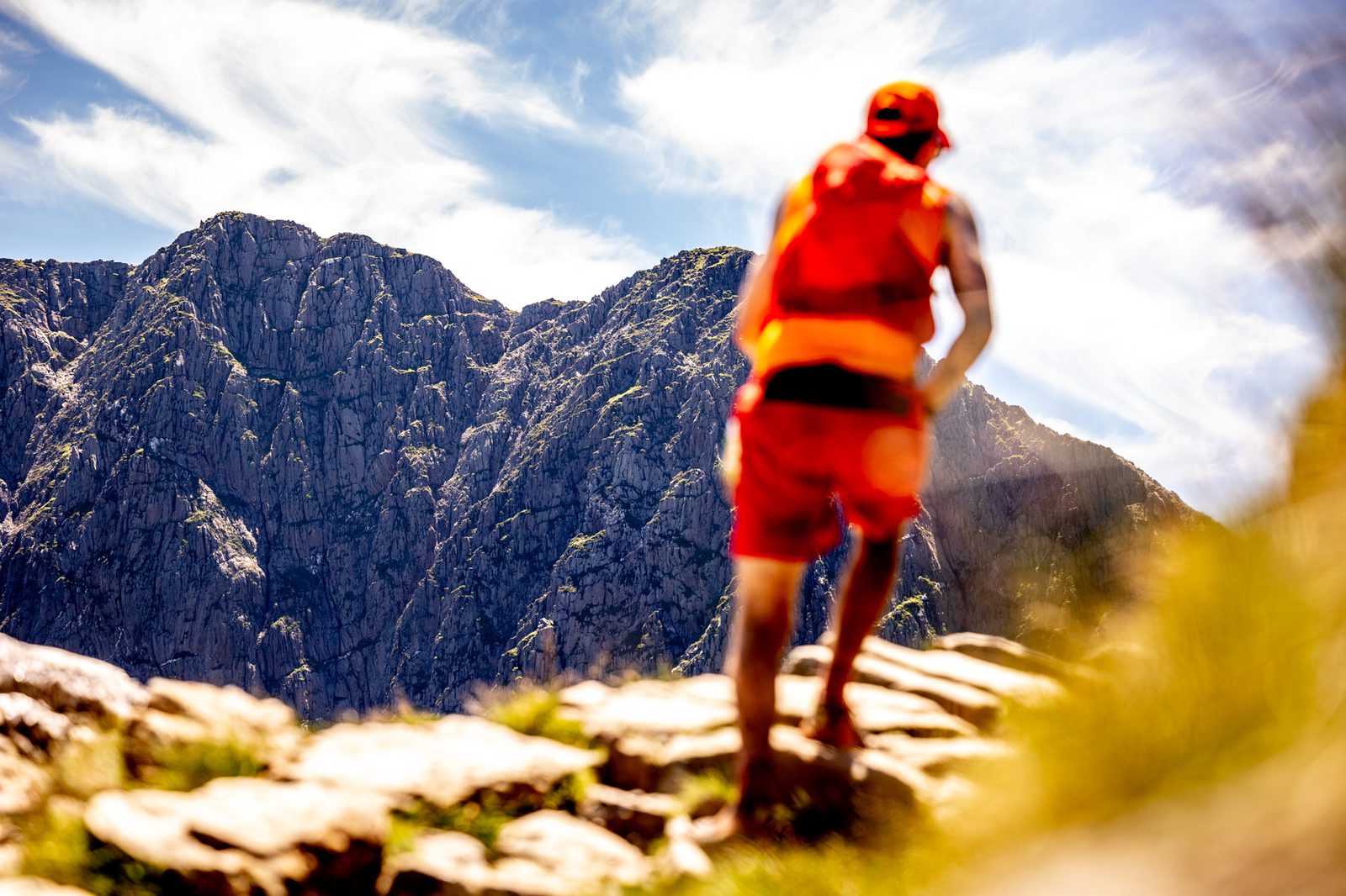 A person wearing bright orange athletic gear and a cap is running or hiking along a rocky path. In the background, there are tall, rugged mountains under a blue sky with wispy clouds. The scene is bathed in sunlight, creating a vibrant atmosphere.