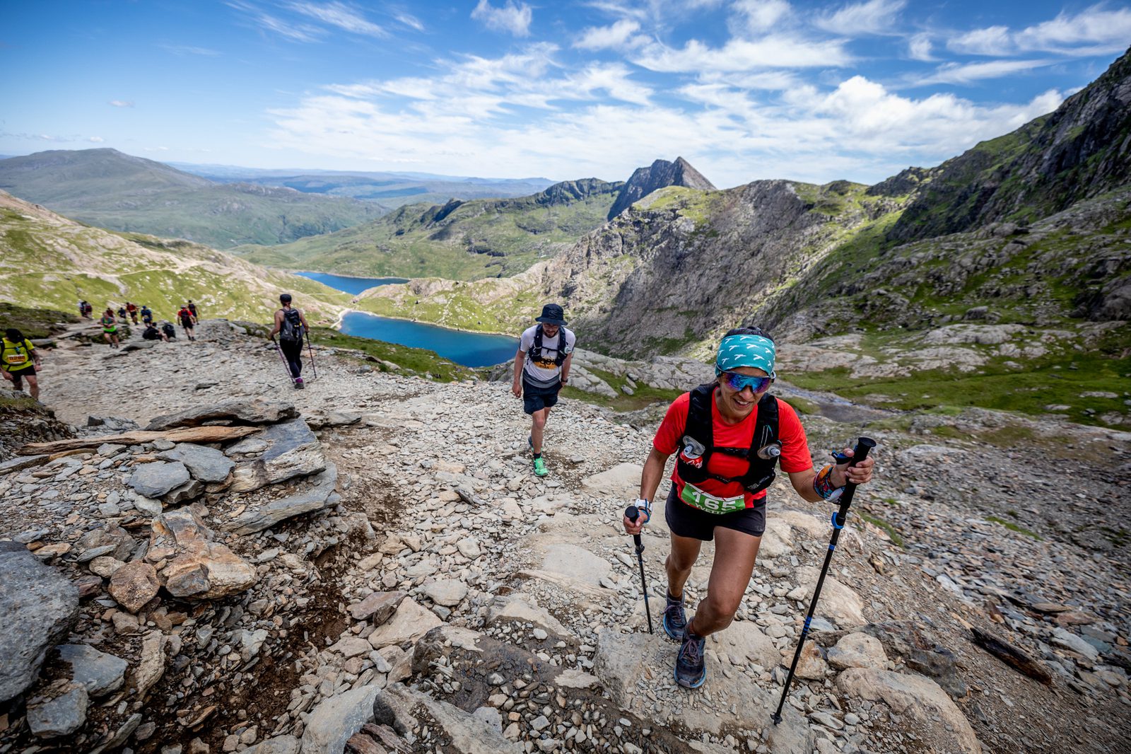 Runners trek up a rocky mountain trail with scenic views of distant peaks and a lake. The sunny day highlights the rugged terrain and blue sky. One runner in the foreground wears a red shirt and uses trekking poles, while other participants follow behind.