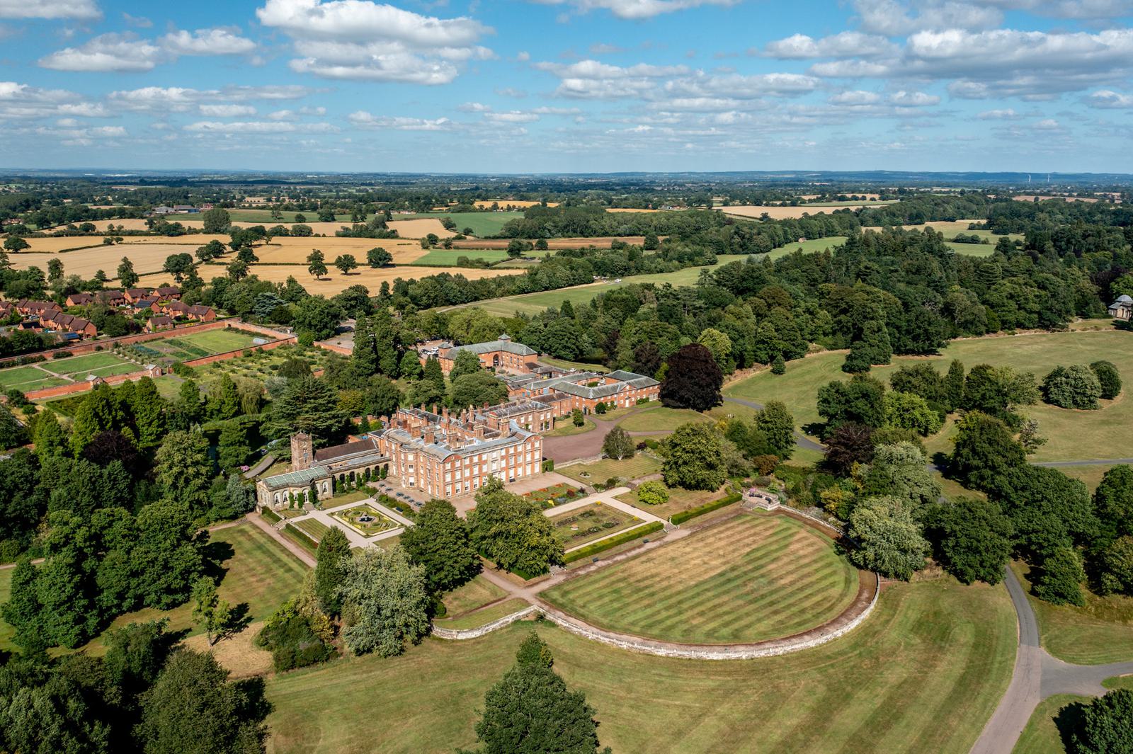 Aerial view of a large, historic estate surrounded by lush greenery and farmland. The estate features a grand, brick mansion with extensive gardens and manicured lawns. Rolling fields and patches of trees are visible under a partly cloudy sky.