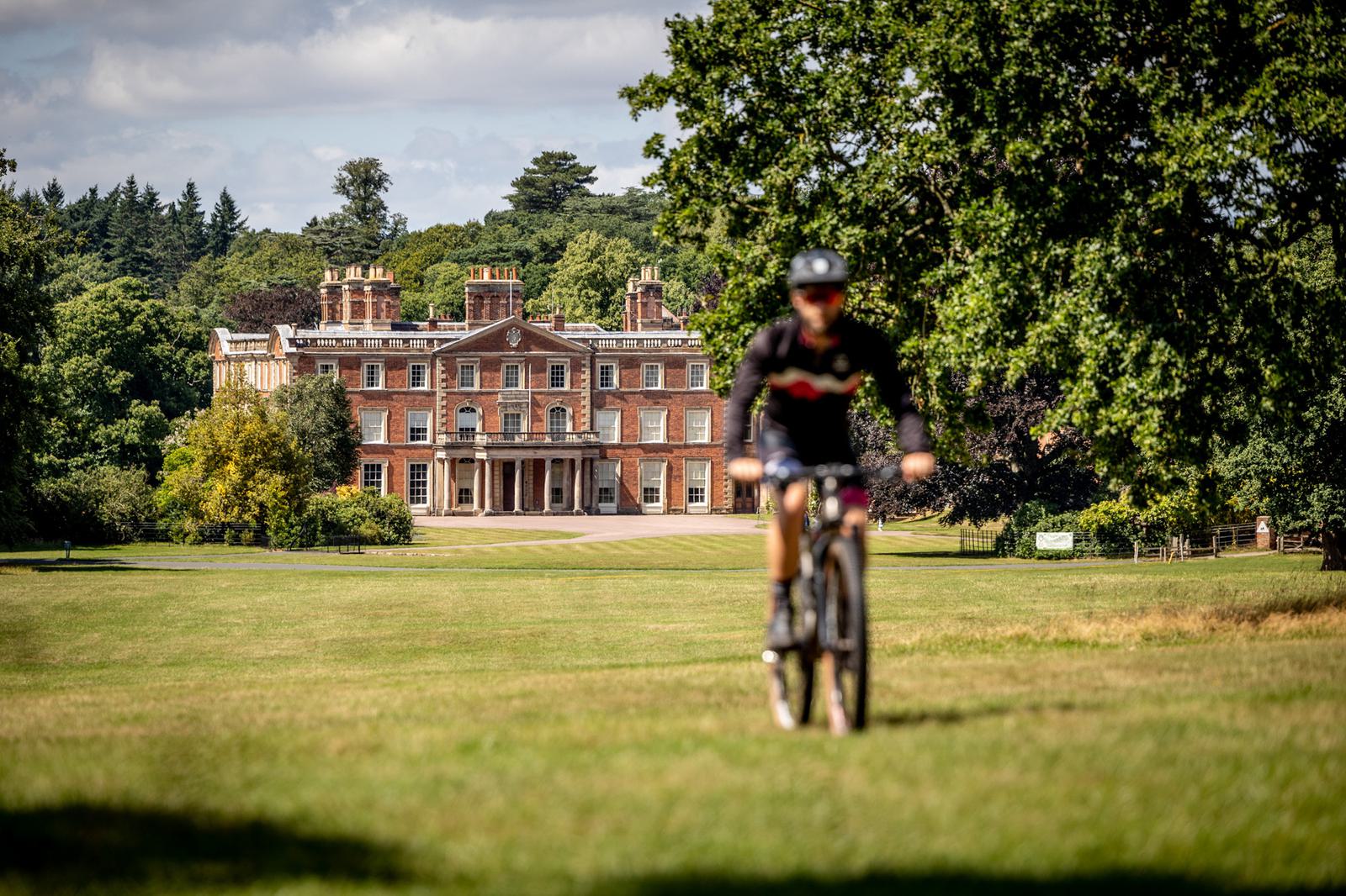 A cyclist rides on a grassy field towards a grand, red-brick manor house with multiple chimneys and large windows, surrounded by lush greenery and trees under a partly cloudy sky. The focus is on the manor house, with the cyclist slightly blurred in the foreground.