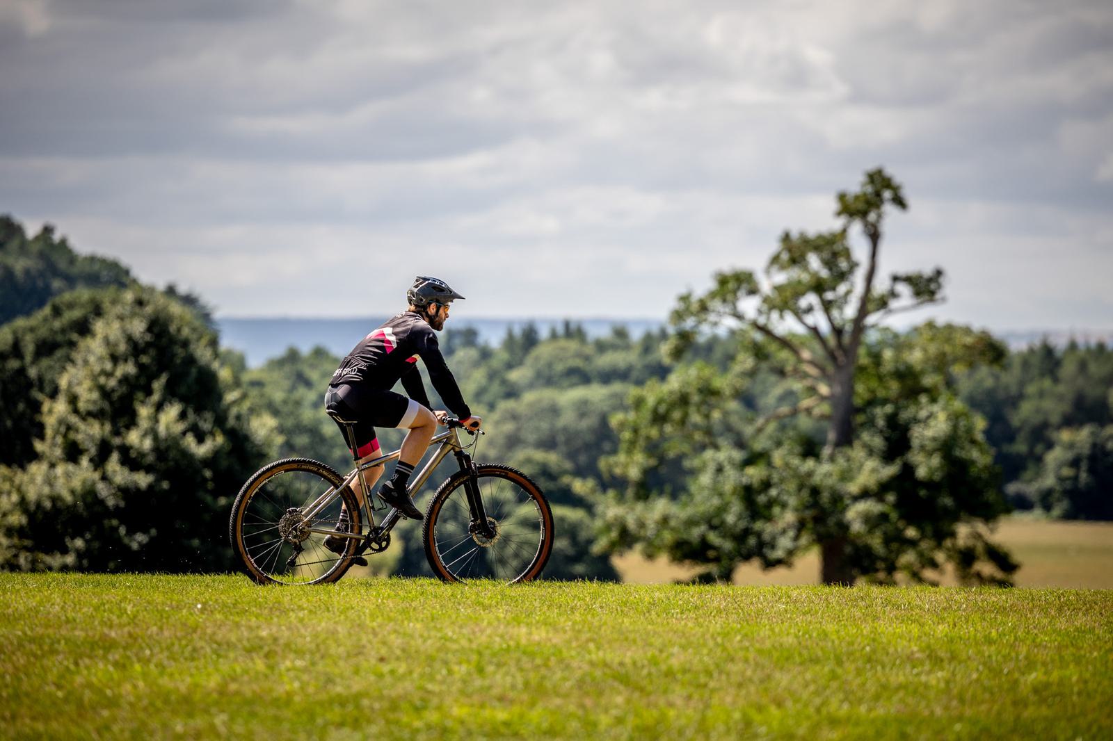 A person wearing a helmet and cycling attire rides a bicycle on a grassy field. The background features a tree, more greenery, and a cloudy sky.