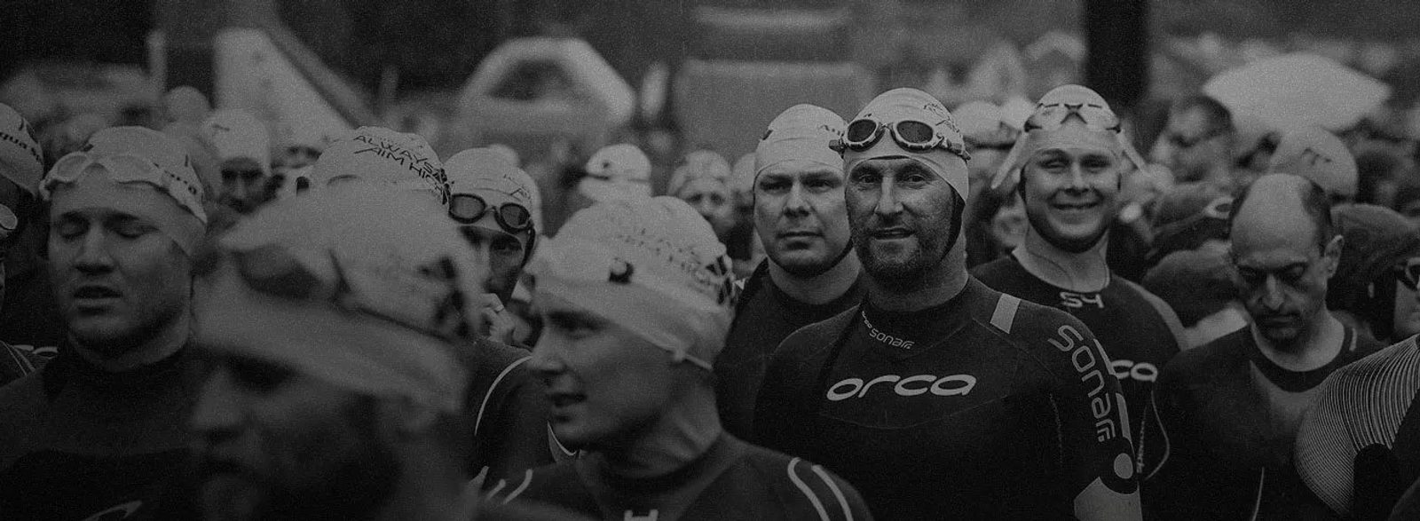 A large group of people wearing swimming caps and wetsuits with goggles on their heads, standing closely together, likely at the start of a swimming or triathlon event. The image is in black and white, showing a mixture of focused and smiling expressions.