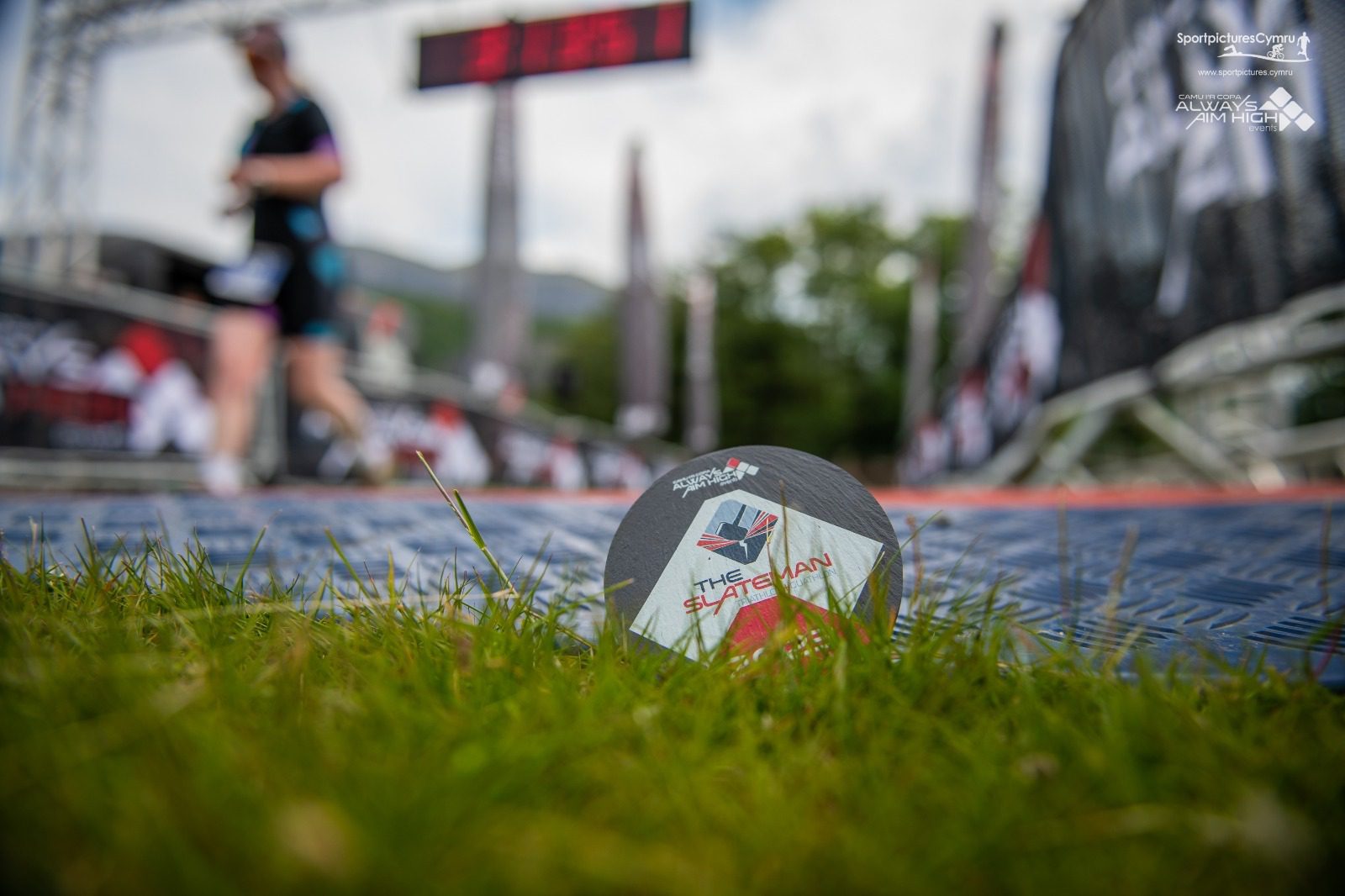 A close-up of a triathlon medal lying on grass, with a blurry athlete and race finish line in the background. The medal displays the event's name, "THE FLATMAN," and a logo with the words "SPORDELICER EVENTS" and "ALWAYS AN ATHLETE" in the top corners.