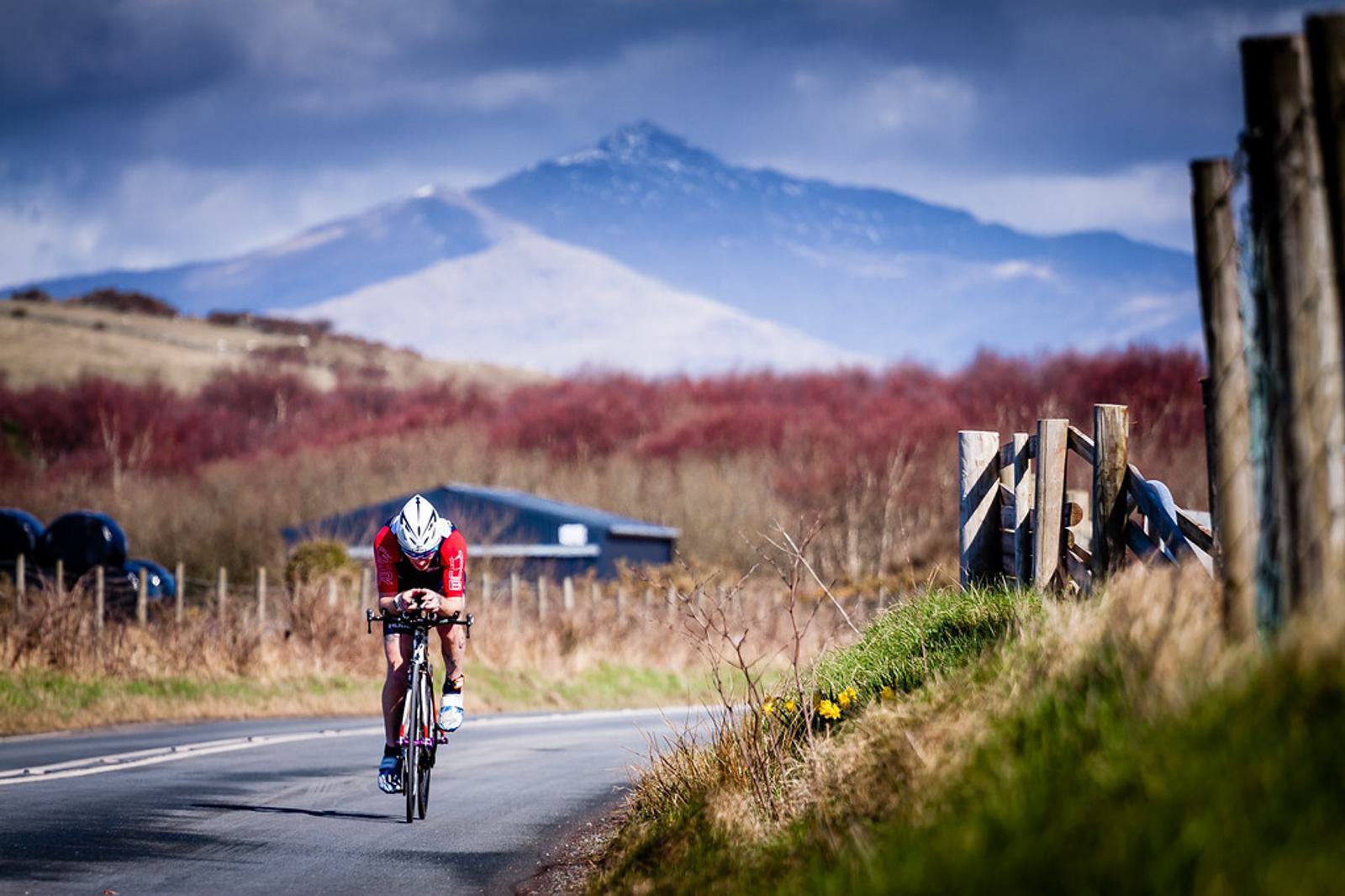 A cyclist wearing a red outfit and white helmet rides on a rural road with a scenic mountain view in the background. The landscape features fields, wooden fences, and a partly cloudy sky, creating a picturesque and serene outdoor setting.