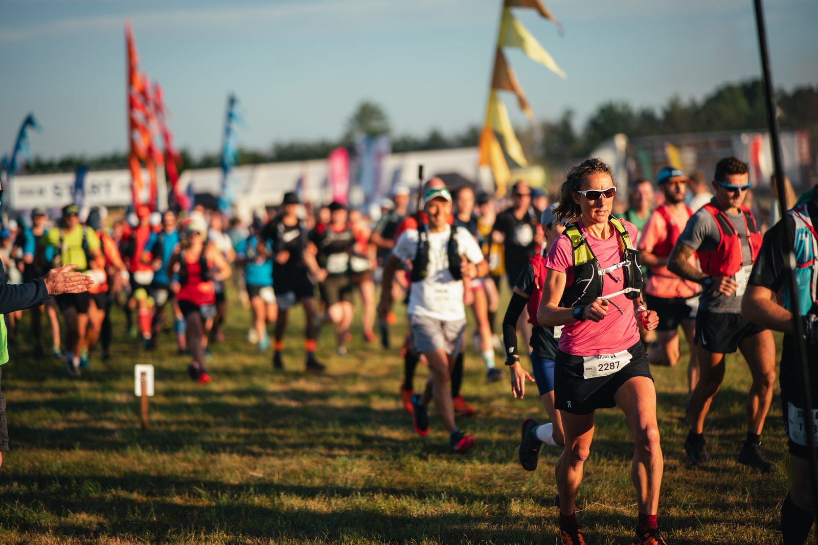A group of runners participates in an outdoor race, with many wearing running bibs and gear. The scene is vibrant, with colorful flags and a clear sky in the background. The grass underfoot indicates a natural, outdoor setting. The runners appear focused and determined.