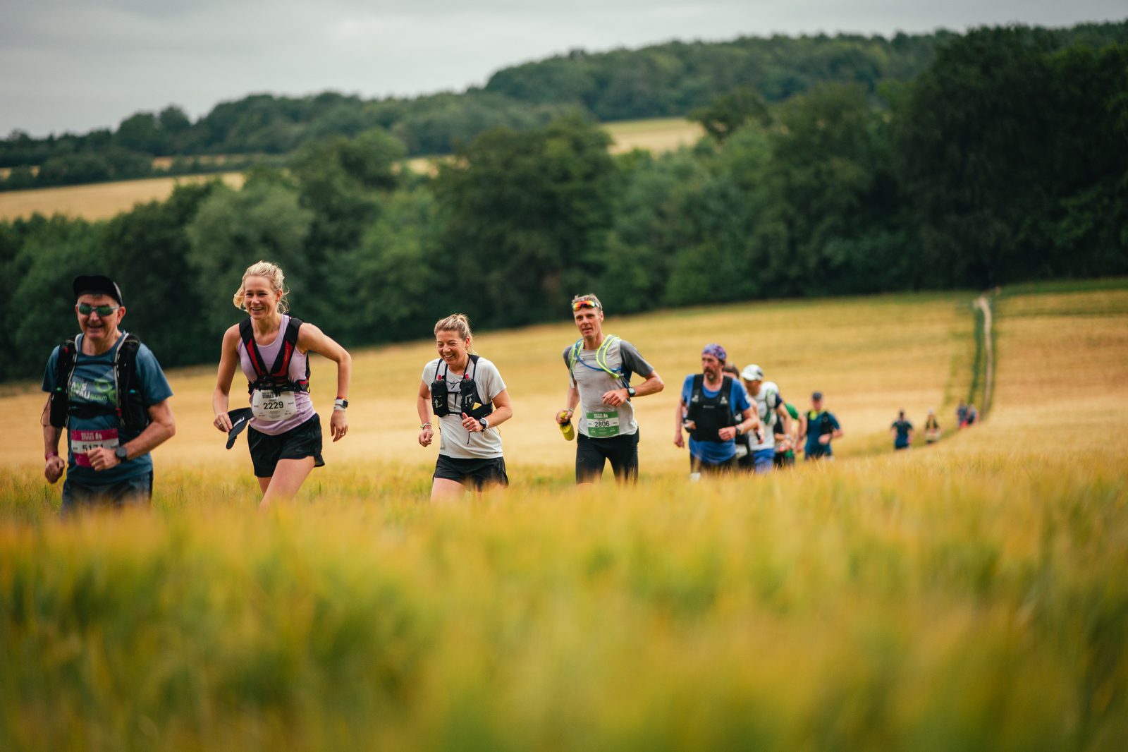 A group of runners participates in an outdoor trail race, traversing a grassy field with trees and hills in the background. They wear athletic gear and race bibs, some smiling as they run. The scene is sunny with a partly cloudy sky.