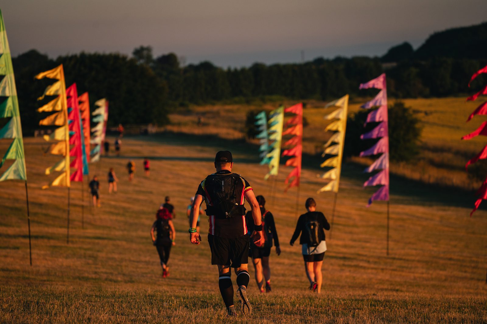 A group of people is running or walking along a grassy path lined with colorful, triangular flags on poles. The sun is low, casting a warm, golden light over the scene. Trees and fields are visible in the background, creating a serene outdoor setting.