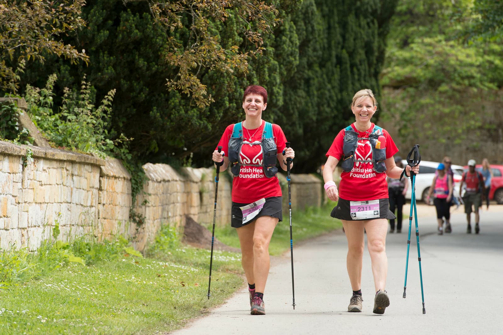 Two women walking along a paved path with trekking poles, both wearing red t-shirts that read "British Heart Foundation." They appear to be participating in an organized trek, surrounded by greenery and a stone wall, smiling and walking energetically.