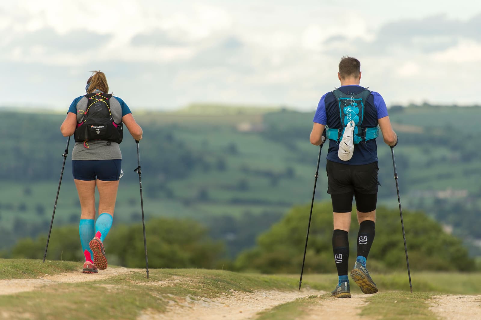 Two people are seen from behind hiking on a dirt trail in an open countryside. They both carry backpacks and use trekking poles, wearing athletic attire. Rolling hills stretch into the background under a partly cloudy sky.
