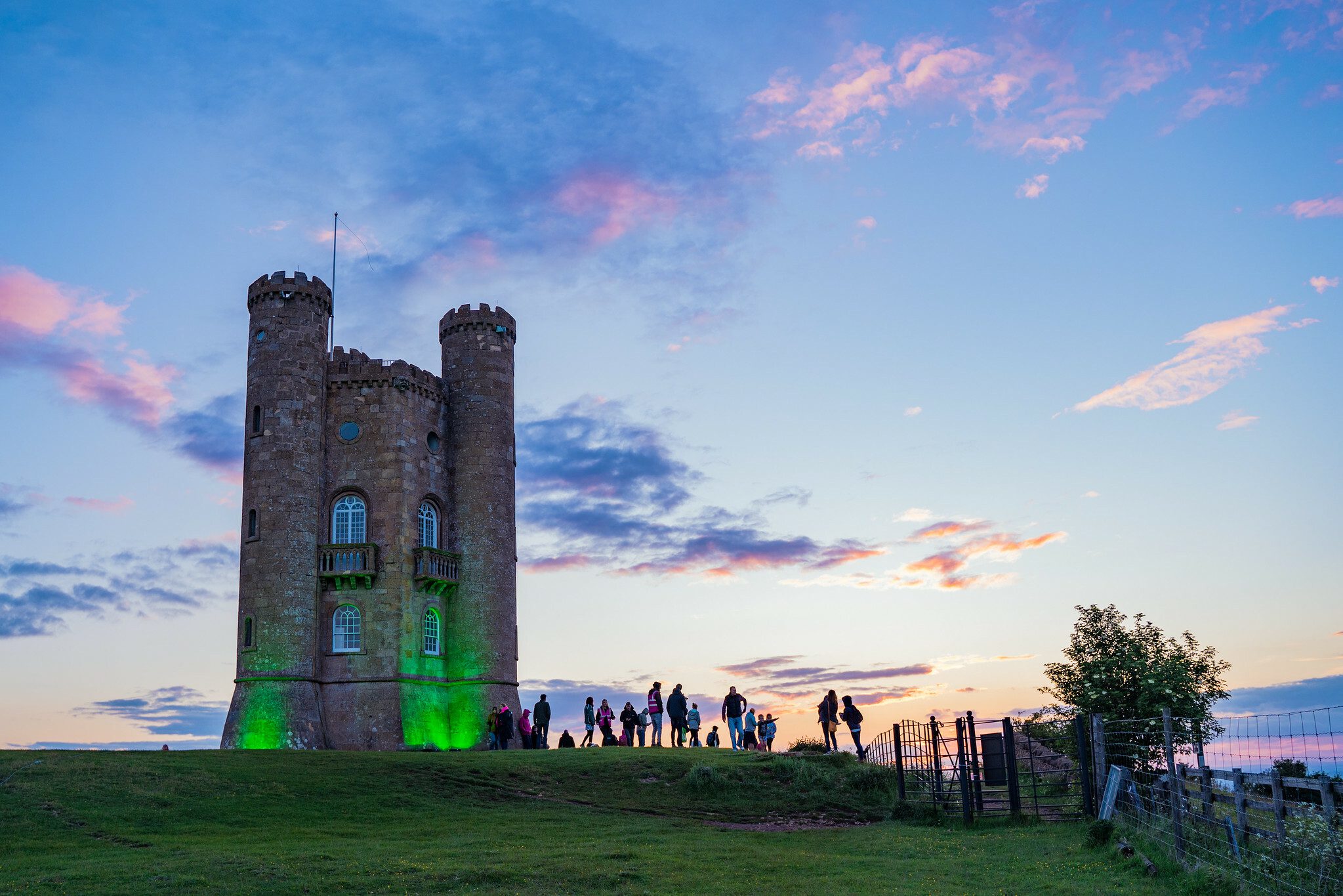 People gather near the illuminated Broadway Tower at sunset. The sky is a mix of pink, blue, and orange hues, creating a picturesque backdrop to the historic tower. Shadows cast by the setting sun add depth to the scene as the group explores the area.