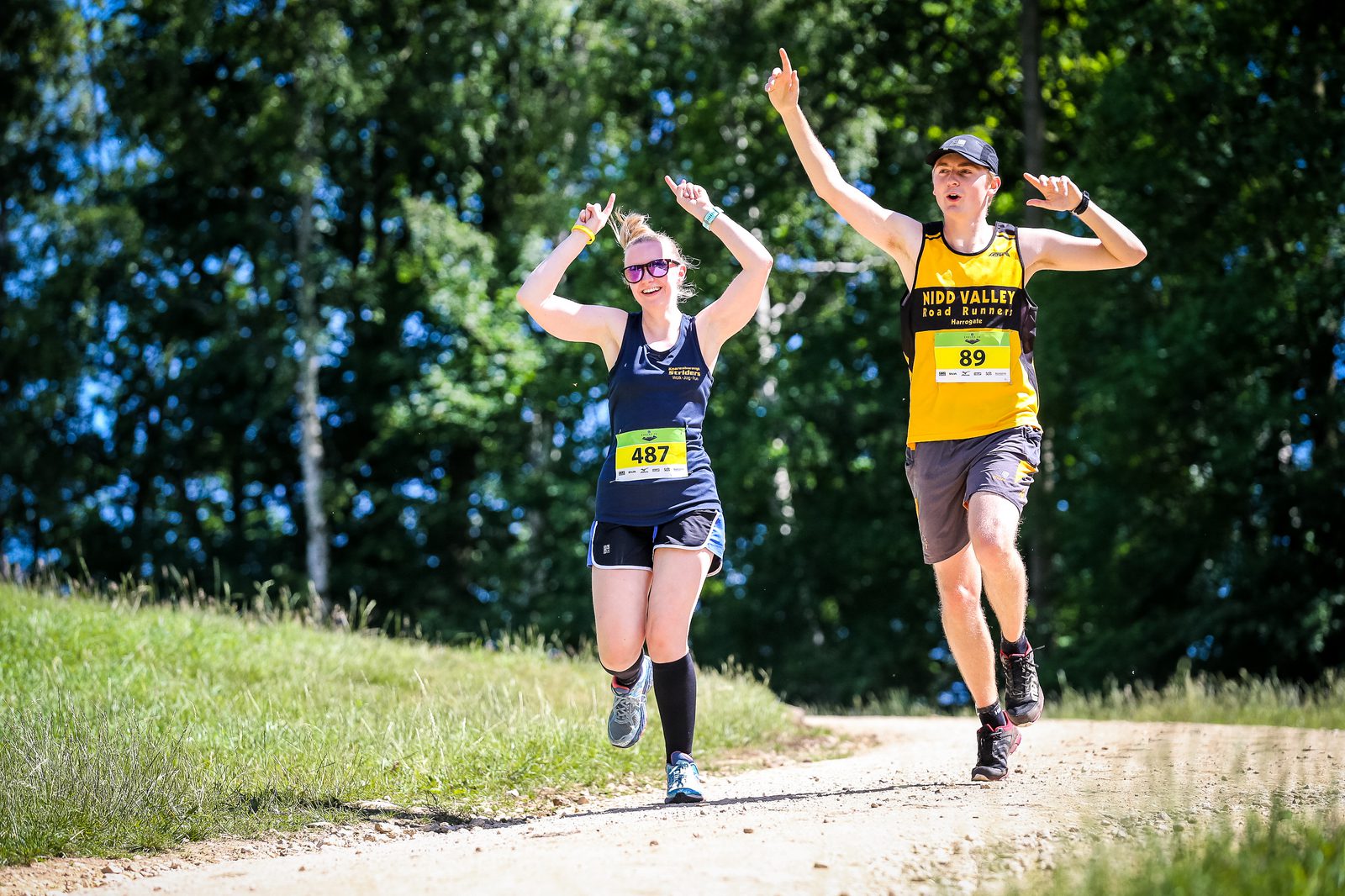 Two runners joyfully raise their arms as they run on a sunny, wooded trail. The woman, in sunglasses and a black outfit, wears a bib number 487. The man, in a yellow and black outfit with a cap, wears a bib number 89. Both are smiling and appear to be celebrating.
