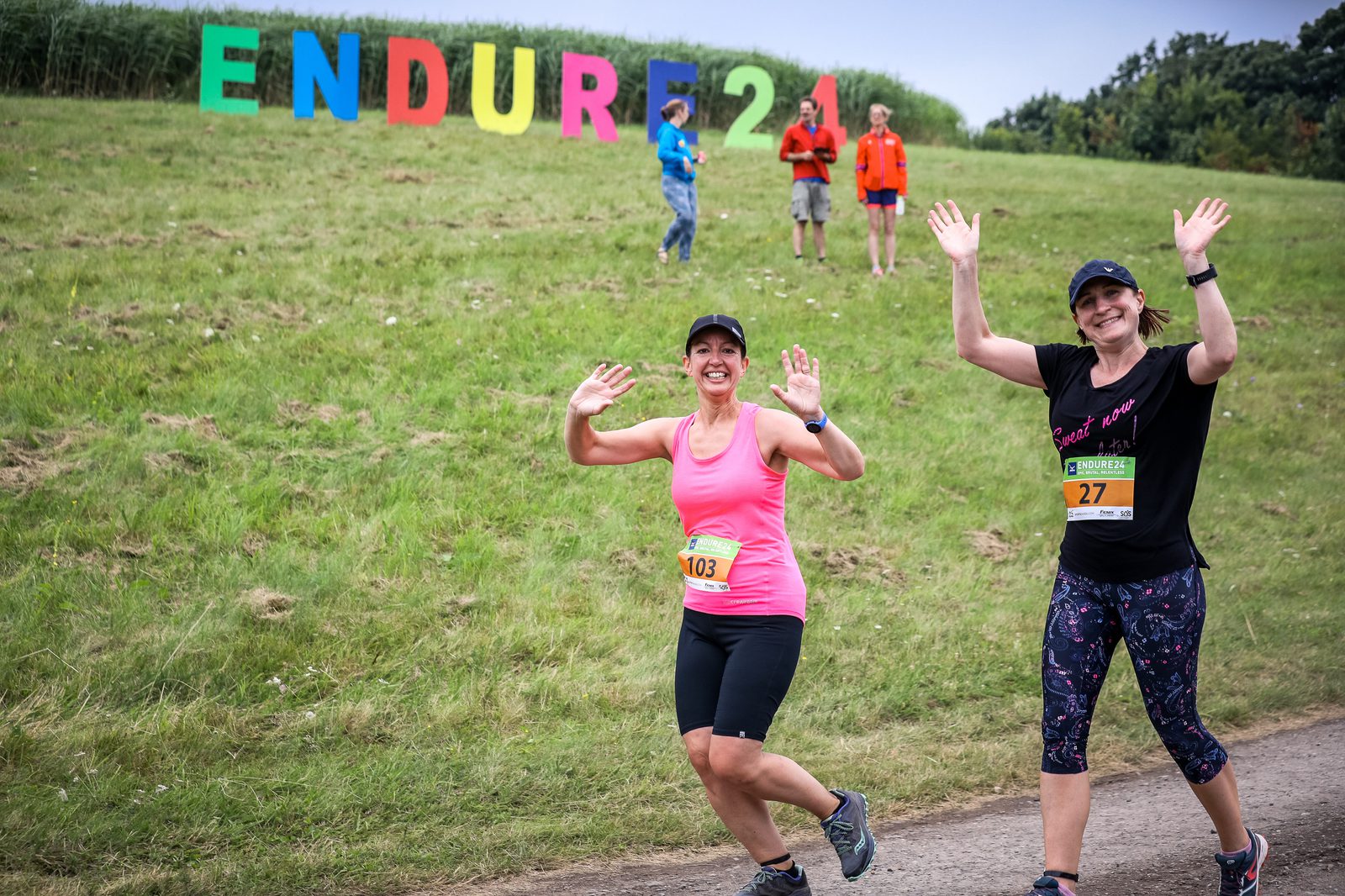 Two women participating in a race wave at the camera, smiling, with colorful "ENDURE 24" letters displayed on a grassy hill behind them. Both wear athletic attire, bib numbers 103 and 27, and appear to be in good spirits. Other participants are visible in the background.