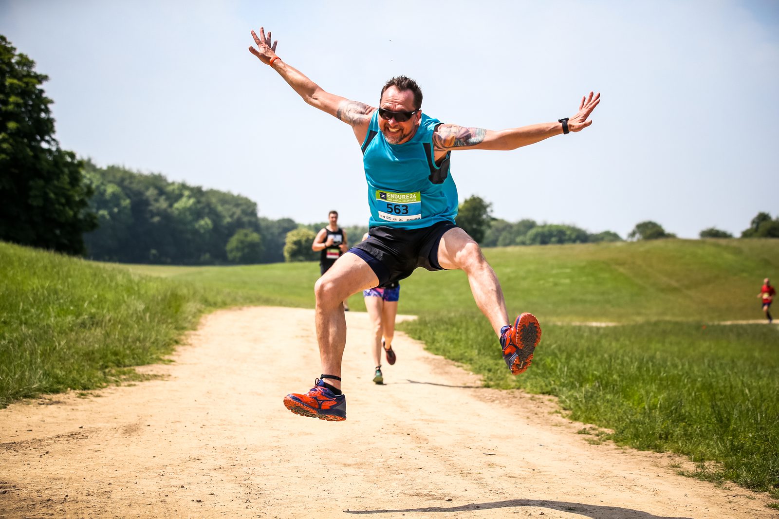 A man wearing sunglasses, a blue tank top, and black shorts is jumping energetically on a dirt path in a park during a race. He has a race bib pinned to his shirt. Other runners can be seen in the background, and the scene is set on a sunny day with green fields and trees.