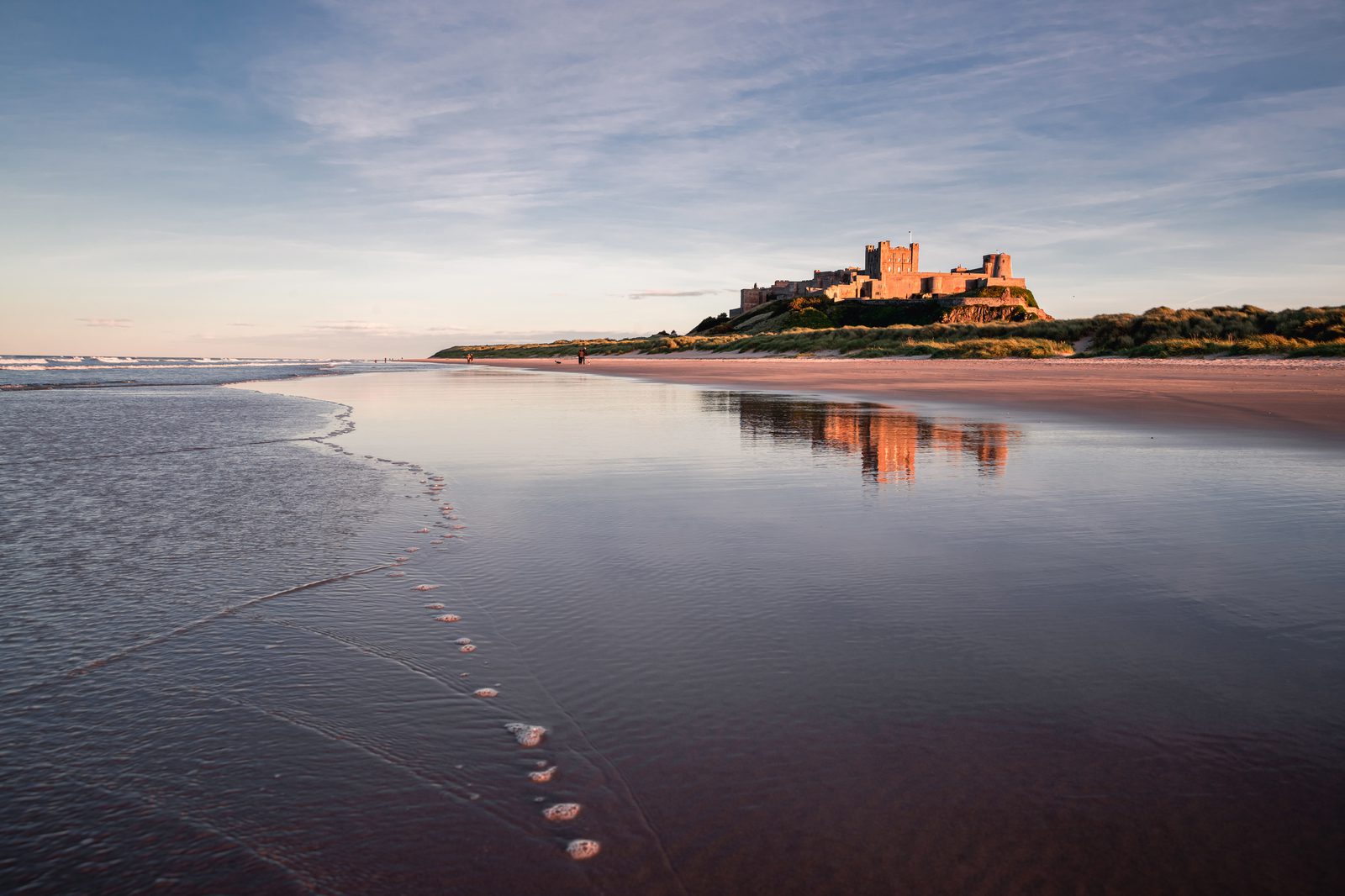 A serene coastal scene at sunrise, featuring a historic castle perched on a low hill in the distance. The castle's reflection shimmers in the calm, shallow water extending across a wide, sandy beach. The sky is mostly clear with soft, warm tones from the rising sun.