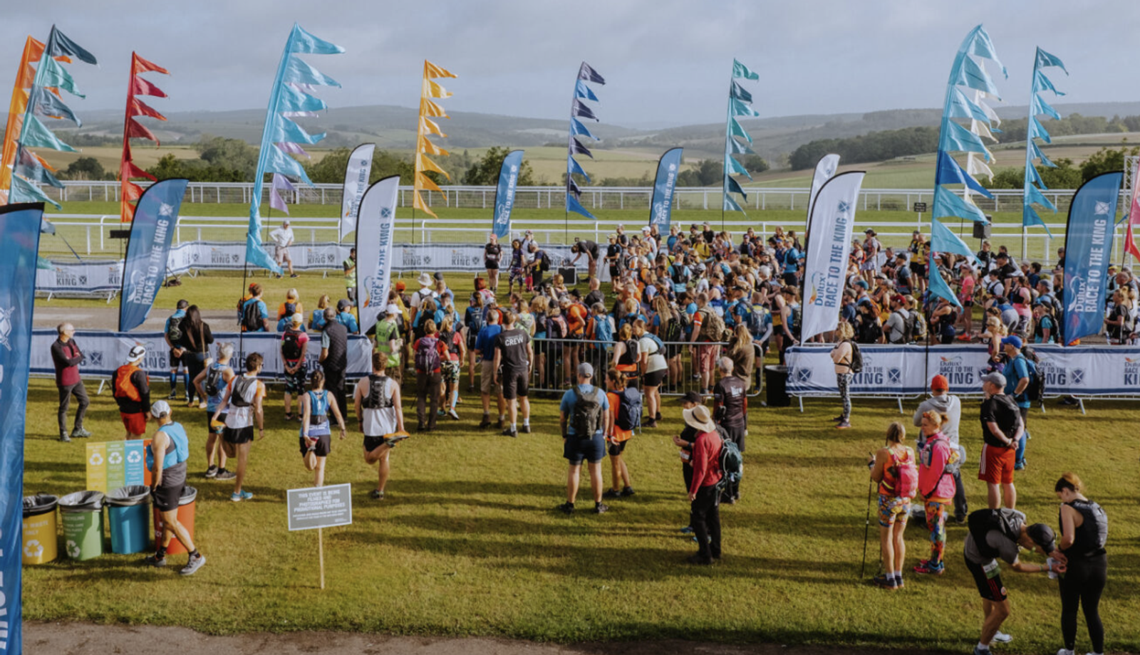 A large group of people, many wearing athletic gear and backpacks, gather at an outdoor event. Colorful flags and banners line the area. A scenic landscape with hills and trees is visible in the background. The sky is partly cloudy.