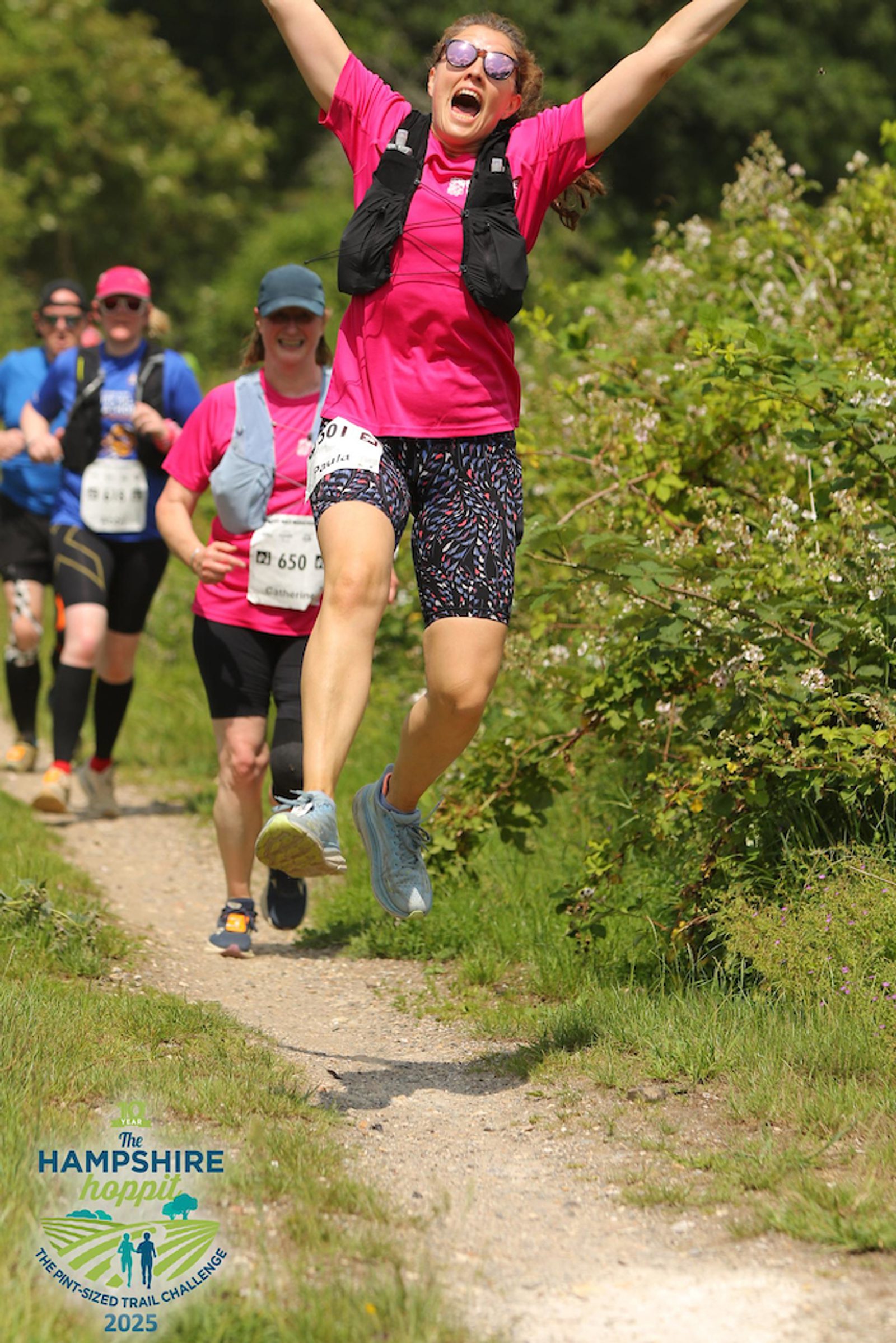 A woman in running gear jumps joyfully in the air on a trail during a race, with other runners behind her. The sun is shining, and greenery surrounds the path. The Hampshire Hoppit race logo appears in the bottom left corner.