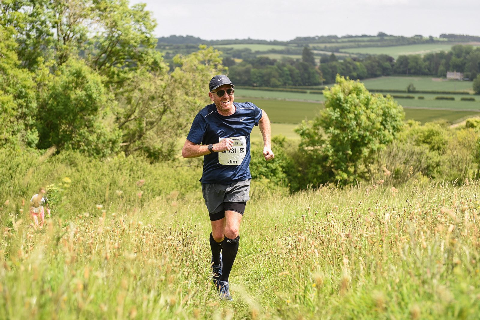 A man wearing a blue shirt, black shorts, running shoes, and a cap is running through a grassy field with a race bib labeled "Jim." The background features lush green trees and rolling hills under a partially cloudy sky.
