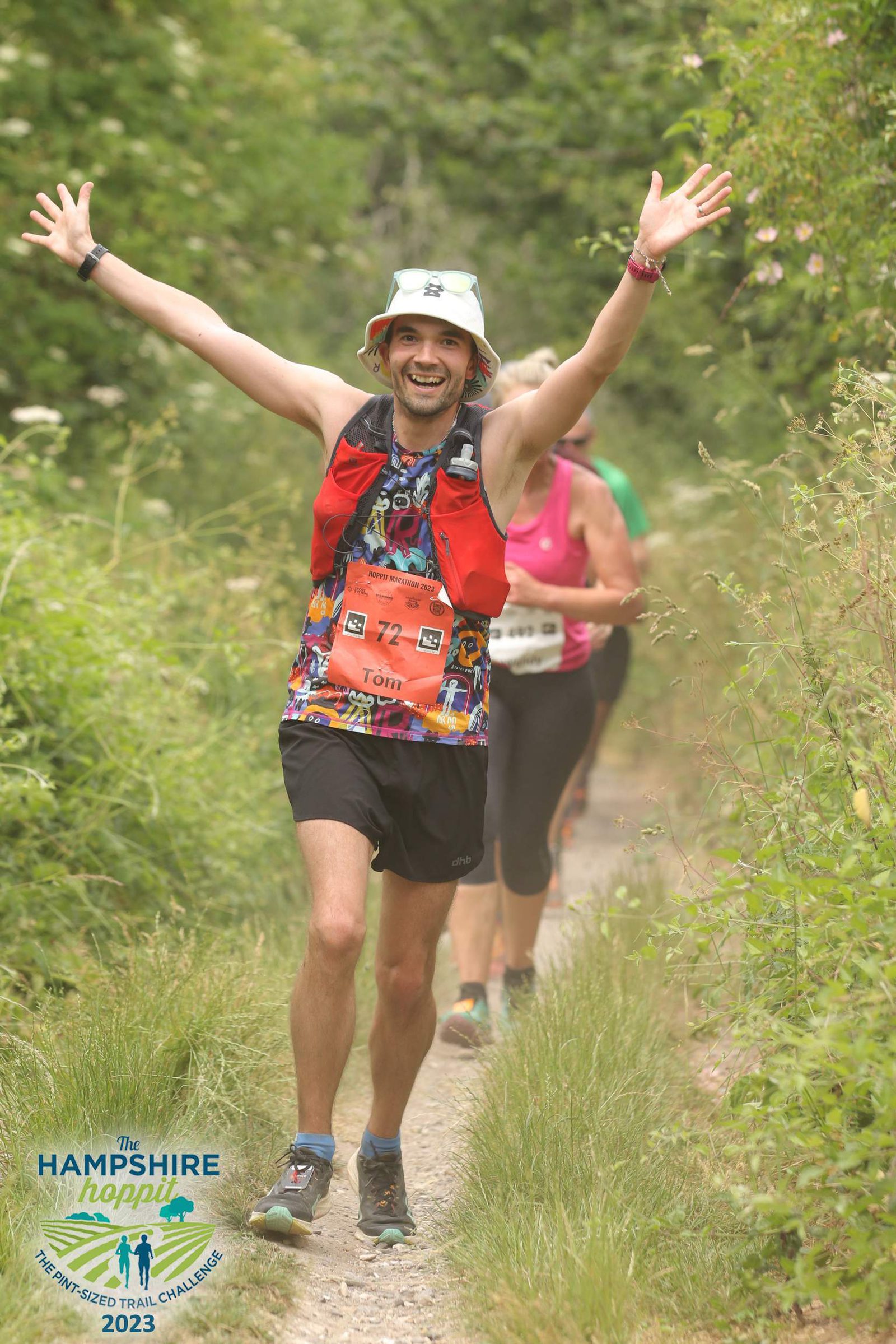 A runner in a bucket hat and athletic gear, including a bright orange bib labeled "Tom," is smiling and raising both arms in celebration while running on a forest trail during The Hampshire Hoppit event. Other runners follow behind, surrounded by lush green foliage.