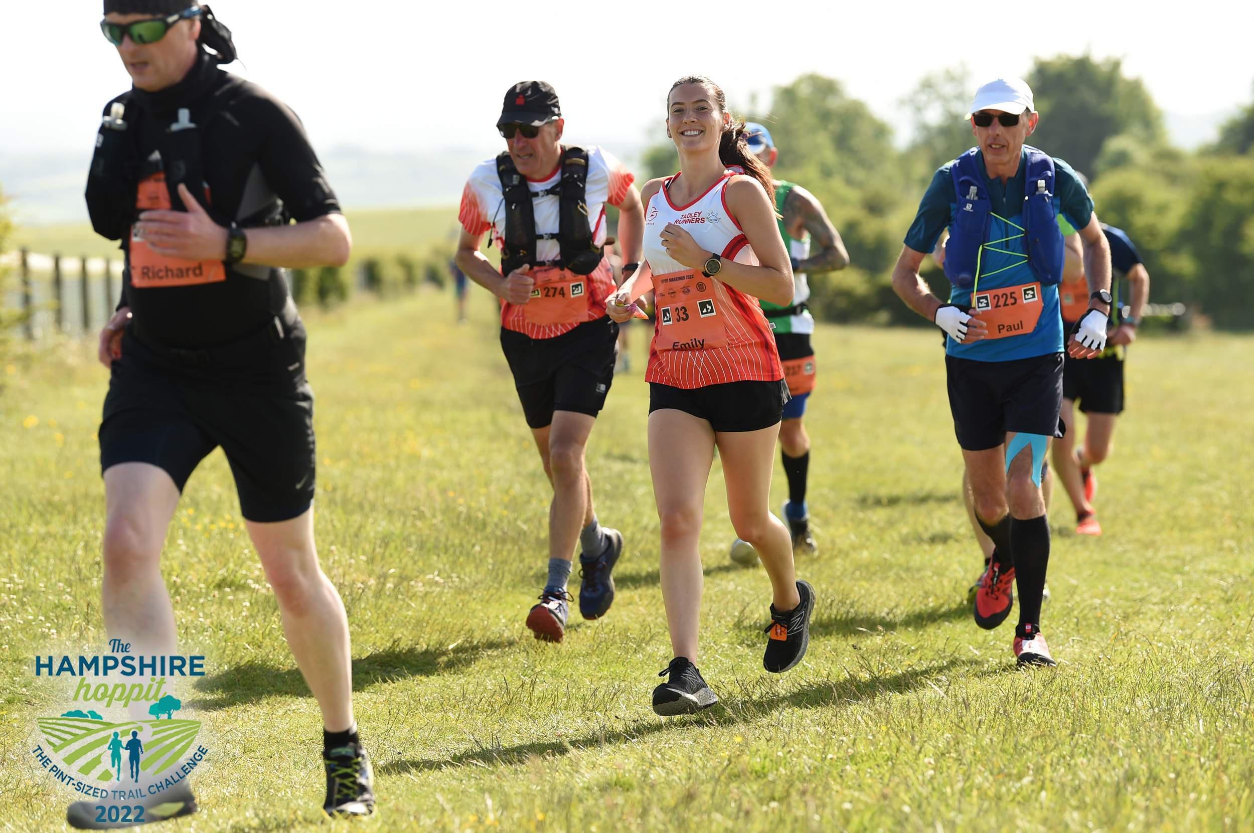 A group of runners are participating in an outdoor marathon event on a grassy field under clear skies. They are wearing athletic gear and bibs with numbers. The image includes an event logo that reads "The Hampshire Hoppit Trail Marathon & Half Marathon 2022.