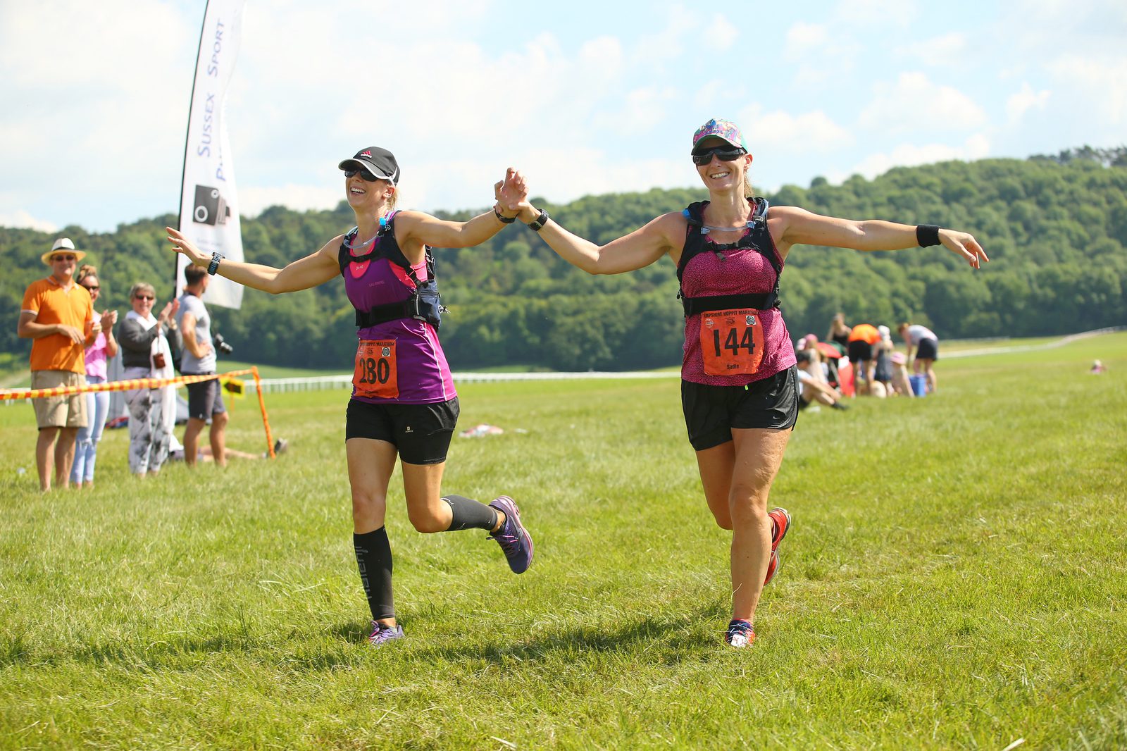Two women in athletic gear, bib numbers 180 and 144, hold hands and smile as they cross the finish line of an outdoor race. Onlookers in the background cheer and take photos. The event appears to take place in a grassy, open area under a clear blue sky.
