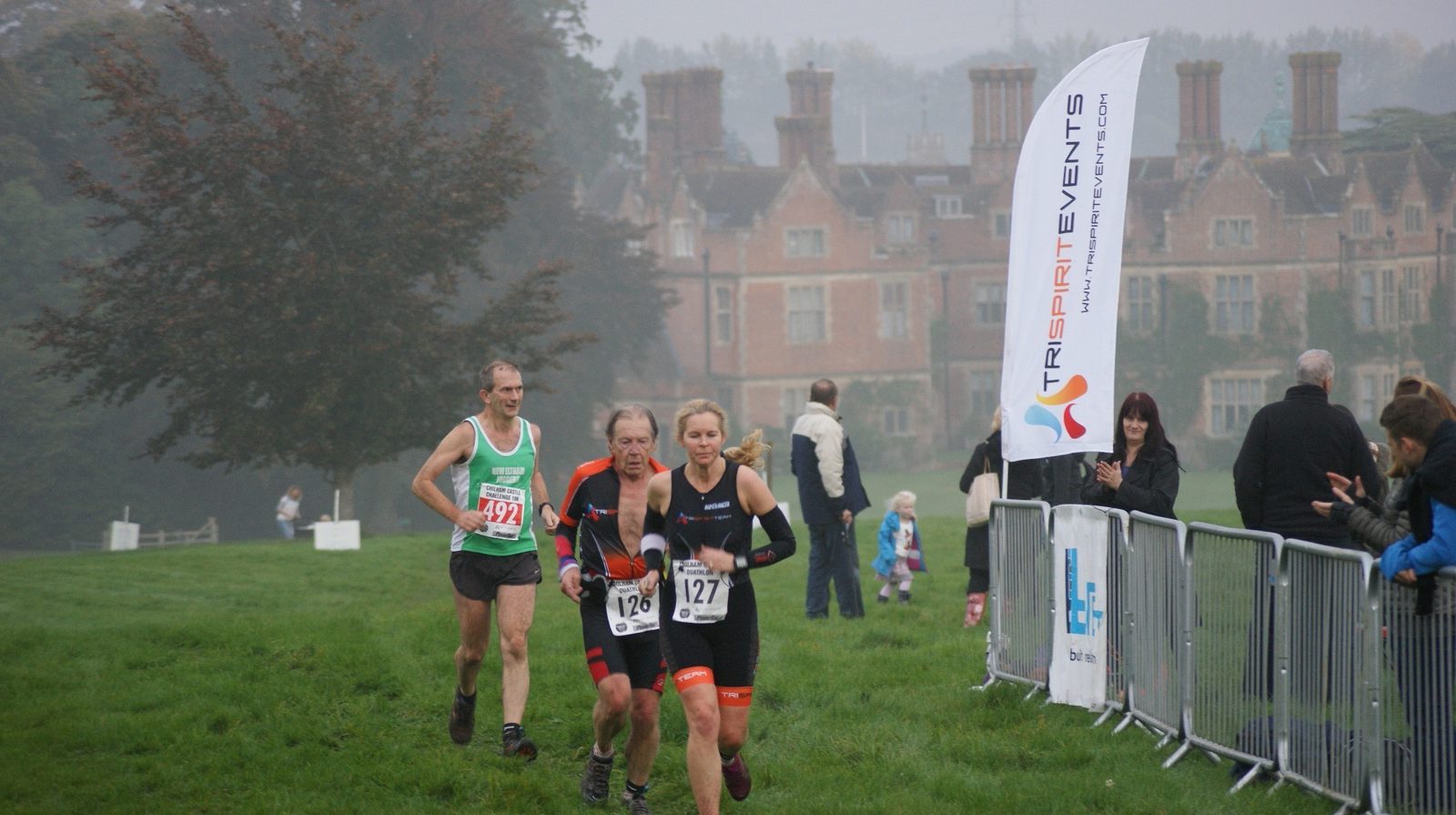 Three athletes running a race on a grassy field with a historic building in the background. Two runners wearing black and orange gear lead, followed by a runner in a green and white uniform. A "Tri Spirit Events" banner and spectators are visible on the right.