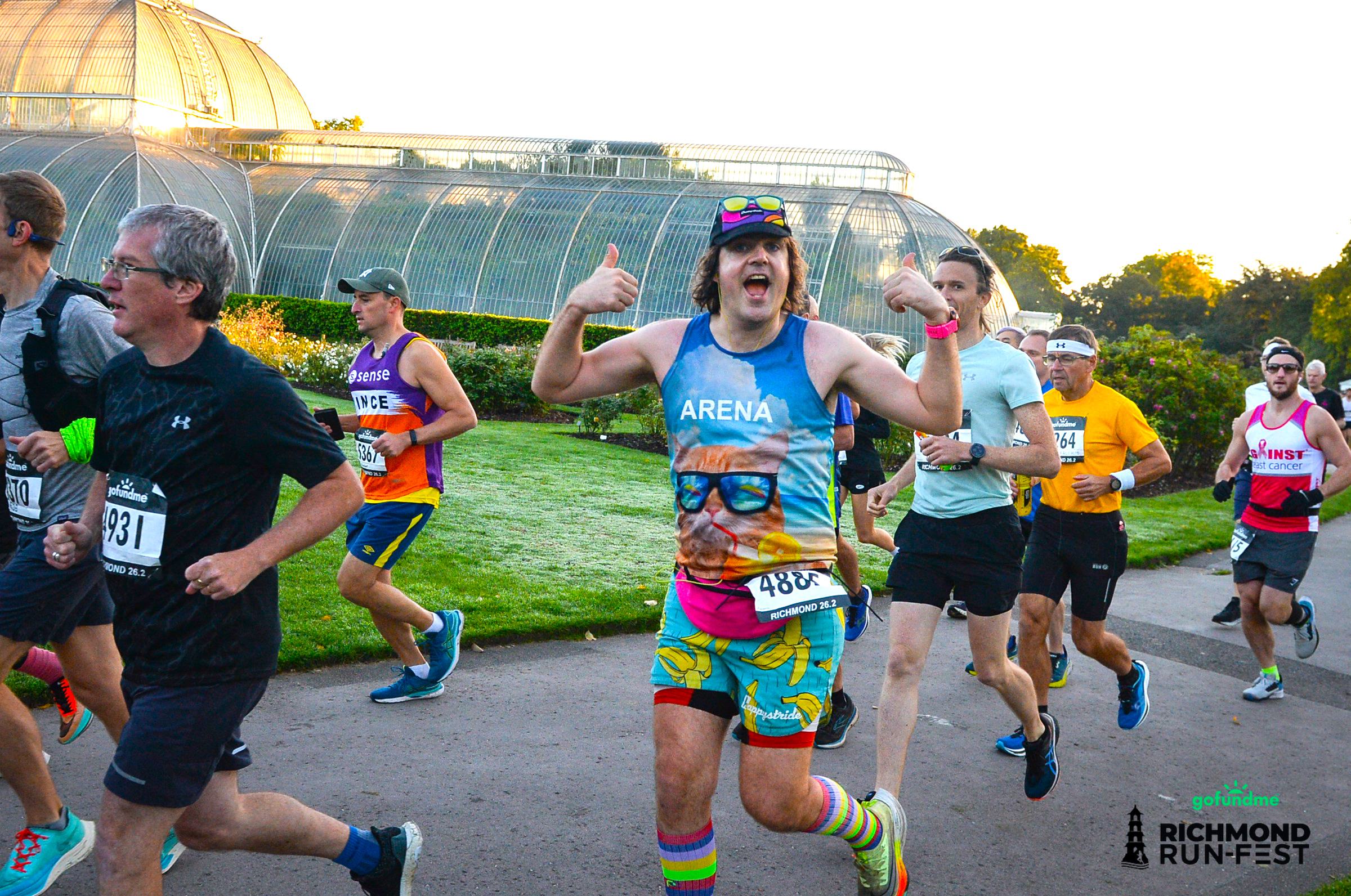 A group of runners participates in a race at the Richmond Run Fest. They run on a path with a large greenhouse in the background. One runner in colorful attire and glasses gives a thumbs up to the camera. Other runners wear race bibs, including numbers 931 and 480.