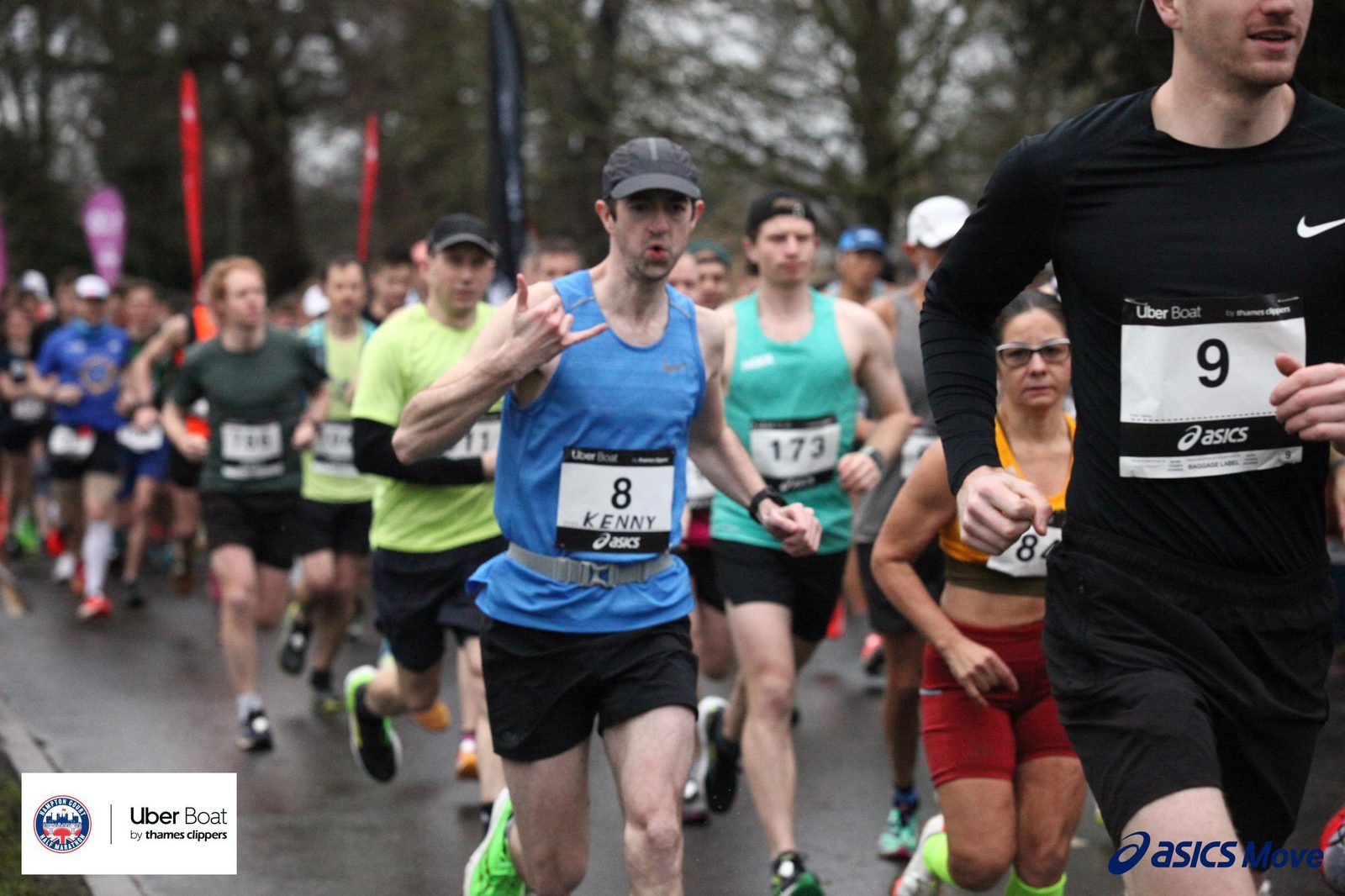 A group of runners, wearing race bibs with numbers, participate in a road race on a wet day. The runner in the foreground wears a blue tank top and black shorts with bib number 8. Other runners are seen behind him wearing a variety of athletic gear. Logos for "Uber Boat" and "ASICS Move" are visible.