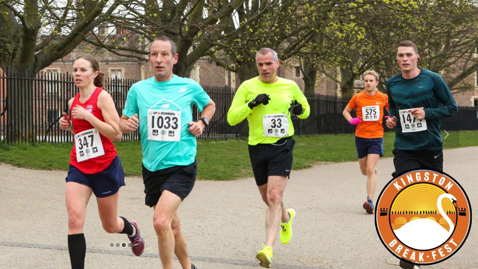 A group of five runners in race attire jog on a park pathway during the Kingston Break-Fest event. Each participant is wearing a numbered bib. Trees with budding leaves can be seen in the background, and the event logo is visible in the corner of the image.