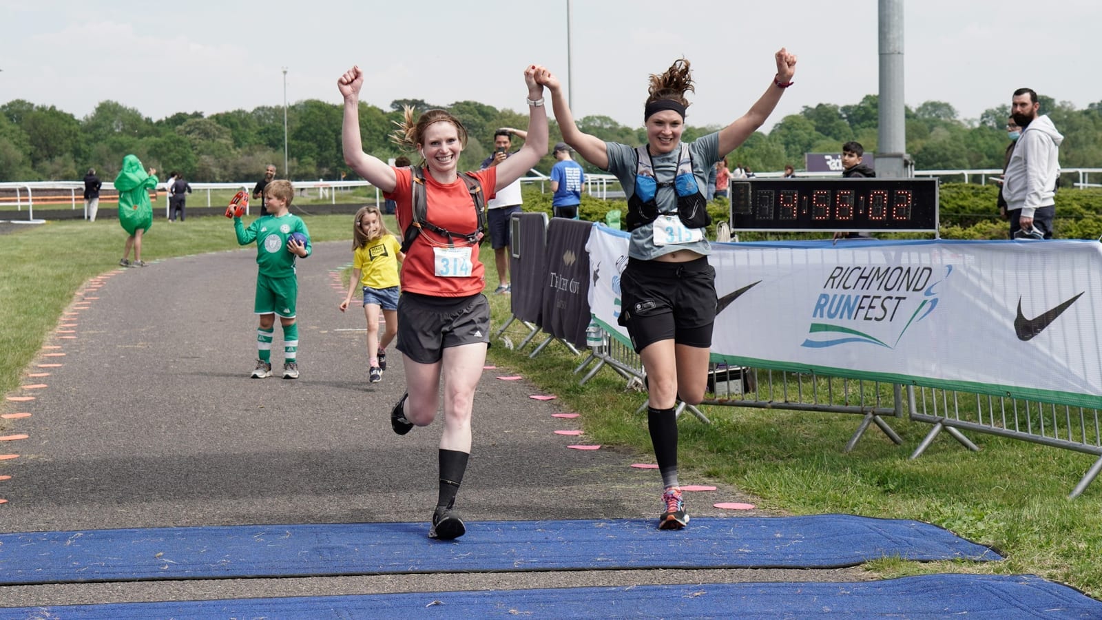 Two runners, one wearing a red shirt (314) and the other a black shirt, cross the finish line of a race at Richmond Runfest, hands raised in victory. Spectators and a digital clock displaying a time of 1:09:51 are visible in the background.