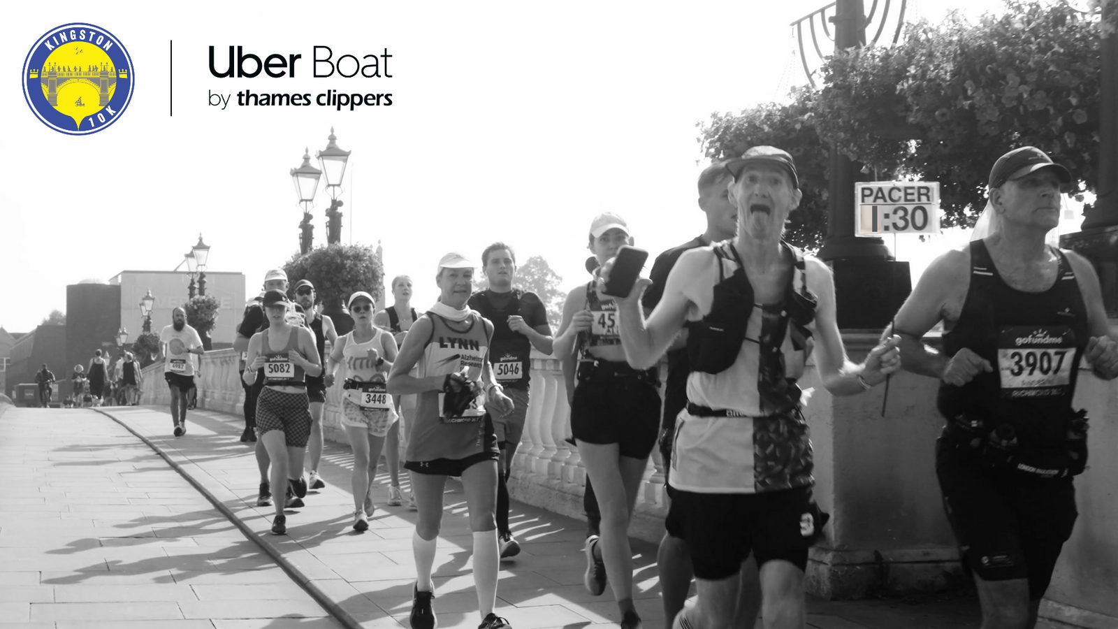 Black and white photo of a group of runners participating in a marathon or race, with some wearing bibs and gear. A "Pacer 1:30" sign is visible among them. The background includes a bridge, street lamps, and the logo of Kingston and Uber Boat by Thames Clippers.