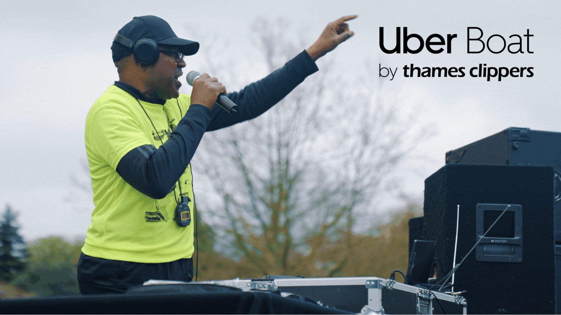 A man in a bright yellow shirt and black cap speaks into a microphone while wearing headphones. He stands in front of audio equipment and points into the distance. The text "Uber Boat by Thames Clippers" is displayed in the upper right corner.