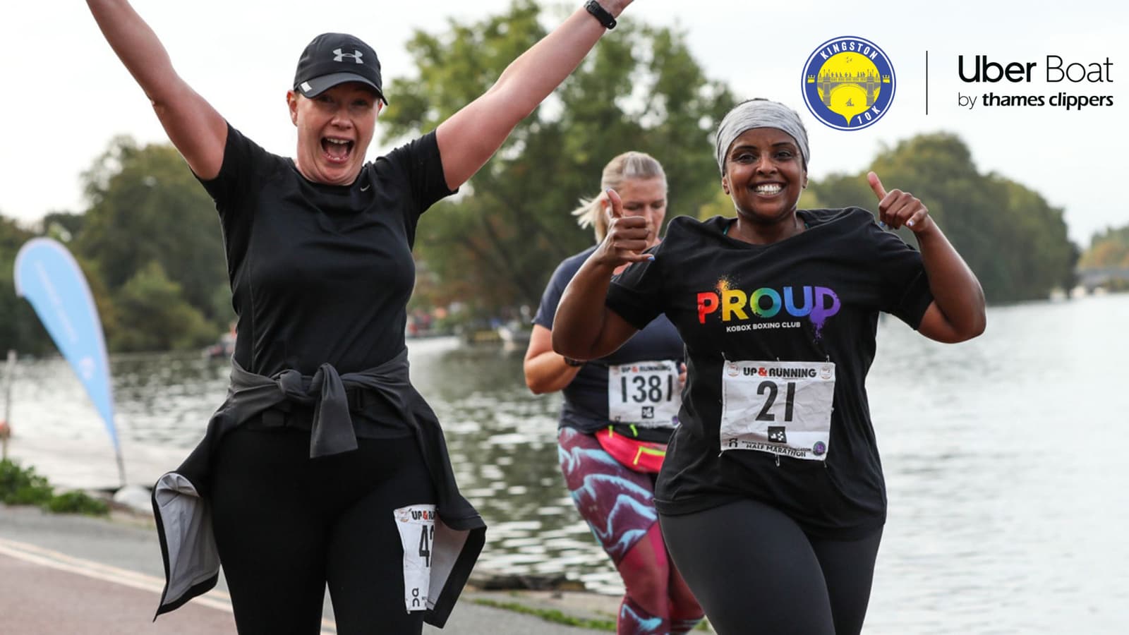Three women are running a race next to a body of water, smiling and cheering. The woman on the left has her arms raised, while the woman on the right gives a thumbs up. They wear race bibs, and the woman on the right has a shirt that reads "PROUD." Logos for Uber Boat by Thames Clippers and Active Training World are in the upper corners.