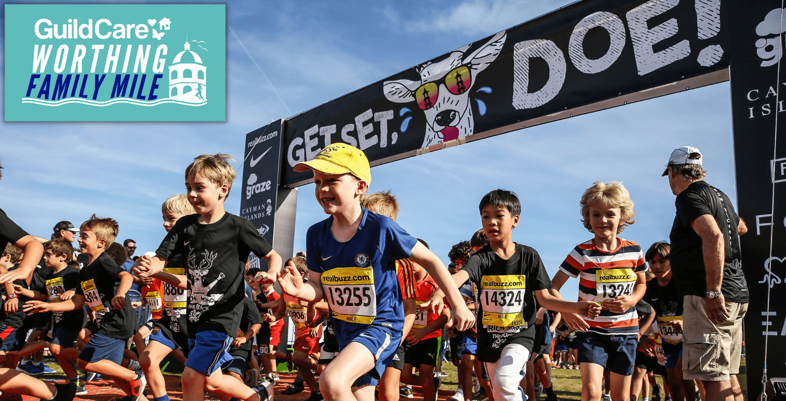 A group of children enthusiastically start running at the GuildCare Worthing Family Mile event. They are seen wearing running outfits and numbered bibs, with a large "Get Set, Go!" banner in the background under a clear sky.