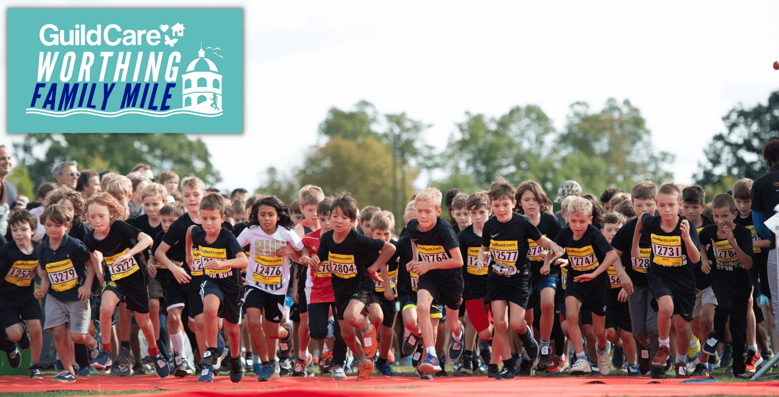 A large group of children wearing black shirts and numbered bibs are energetically starting a race on an outdoor track. In the top left corner, a logo reads "GuildCare Worthing Family Mile" with an image of the Worthing Dome. Trees and a cloudy sky are in the background.