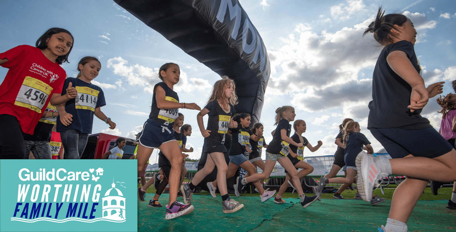 Children are running in a race under an inflatable arch on a partly cloudy day. They wear numbered bibs and athletic clothing. A banner in the bottom left corner reads, "GuildCare Worthing Family Mile" with an illustration of a castle.