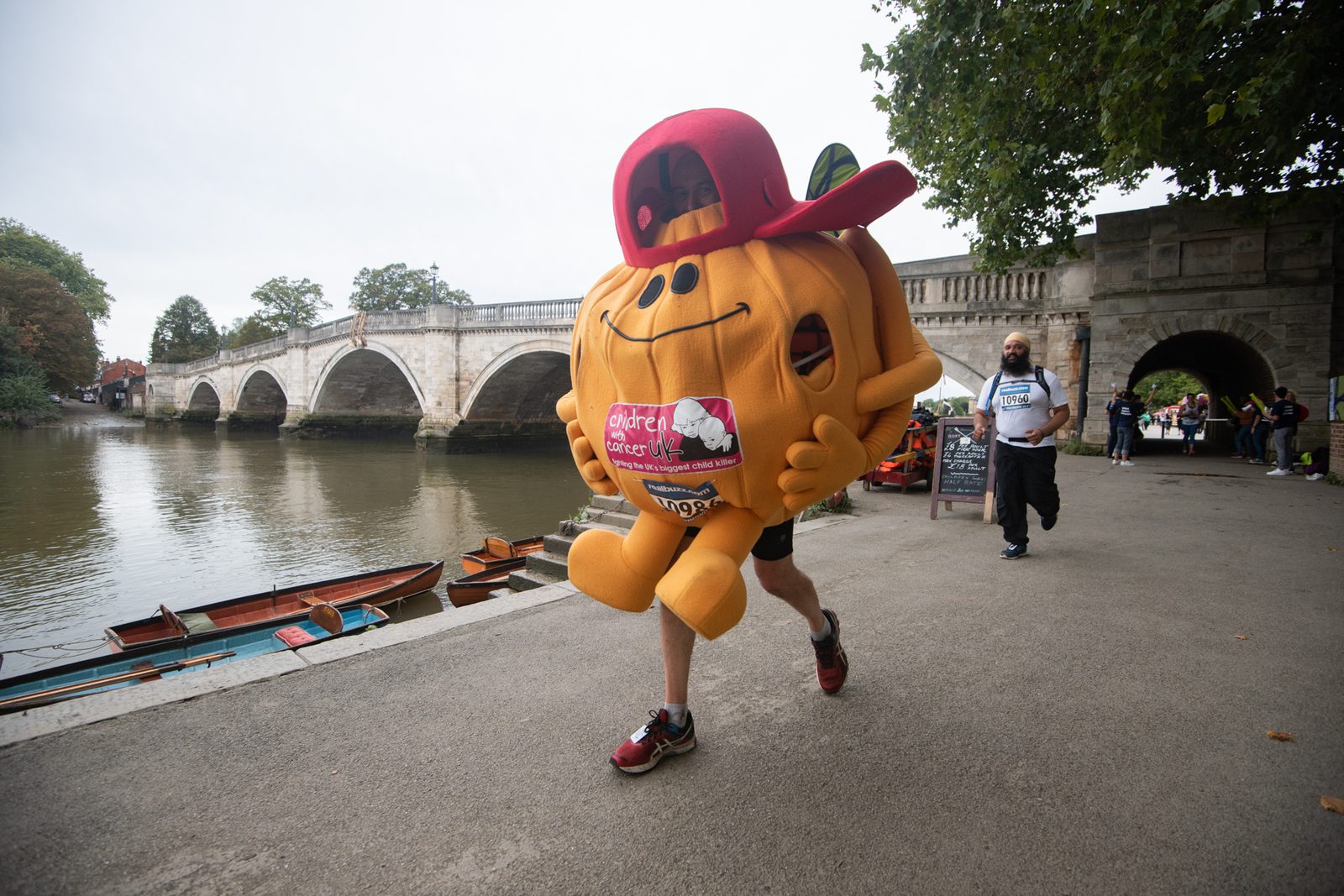 A person dressed in a large pumpkin costume and red cap is running along a riverside path during a fun run event. A bridge spans the river in the background, and a small crowd, including a person pushing a stroller, appears alongside the path.