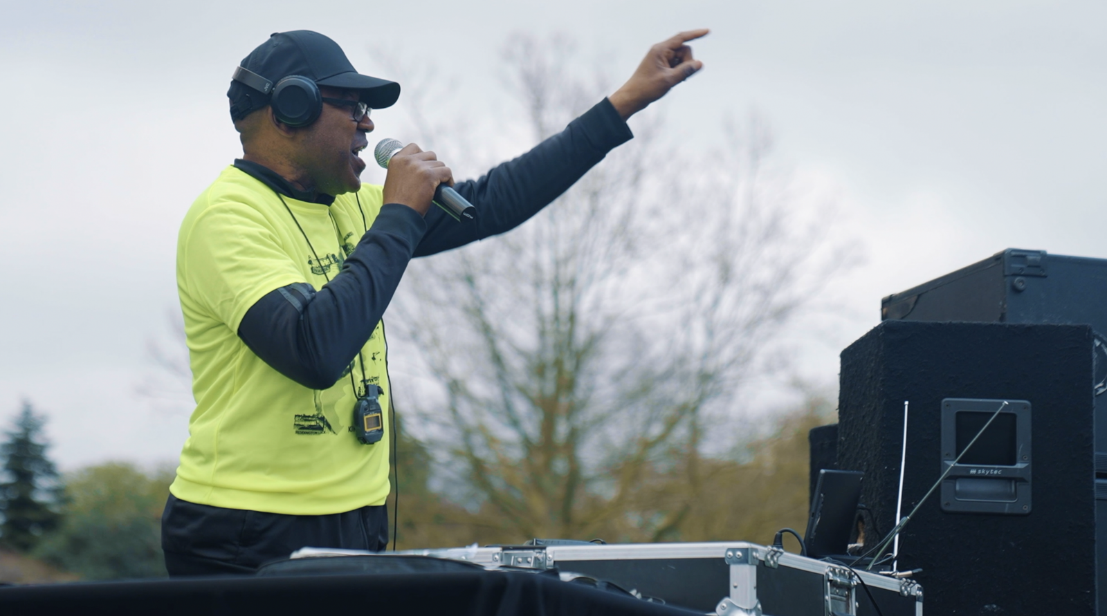 A person wearing a bright yellow shirt, black cap, and sunglasses, holding a microphone and pointing, stands behind DJ equipment. They appear to be speaking or singing into the microphone. There are trees and a cloudy sky in the background.