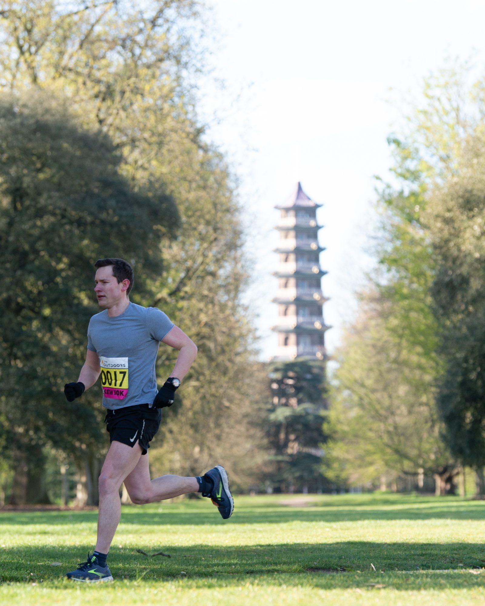 A man wearing a race bib and athletic clothing runs on a grassy path in a park. In the background, a tall pagoda is visible among the trees, suggesting an oriental garden setting. The scene is sunny and vibrant with lush greenery all around.