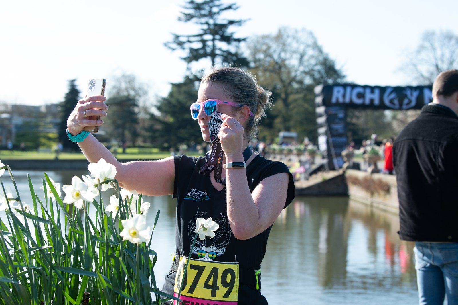 A runner wearing a race number 749 takes a selfie while holding a medal, standing next to a lake with blooming flowers. Trees and race banners are visible in the background. The sun is shining, and the atmosphere appears celebratory.